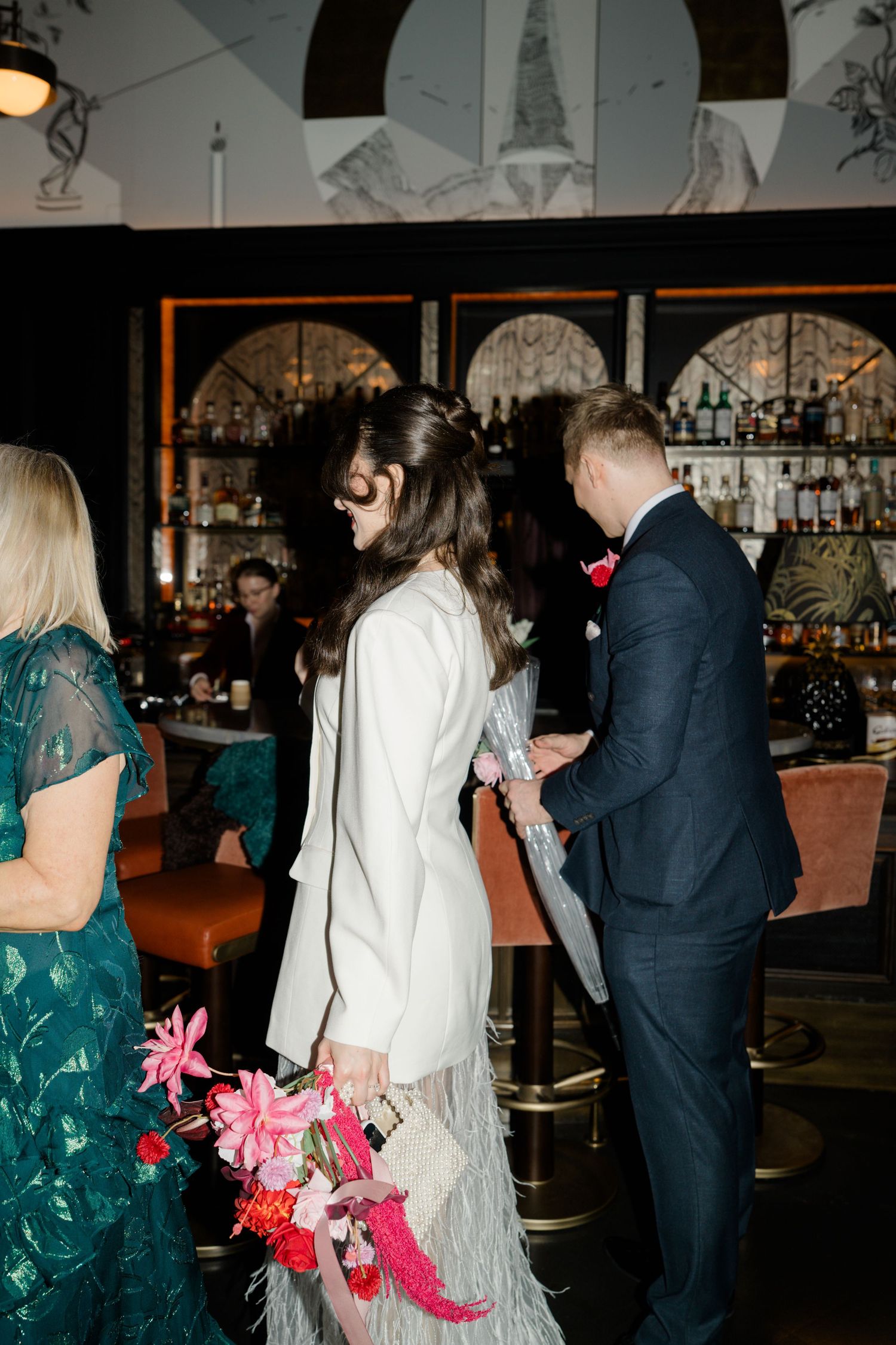 Bride in white feather skirt and groom in navy suit walk through an elegant bar venue.