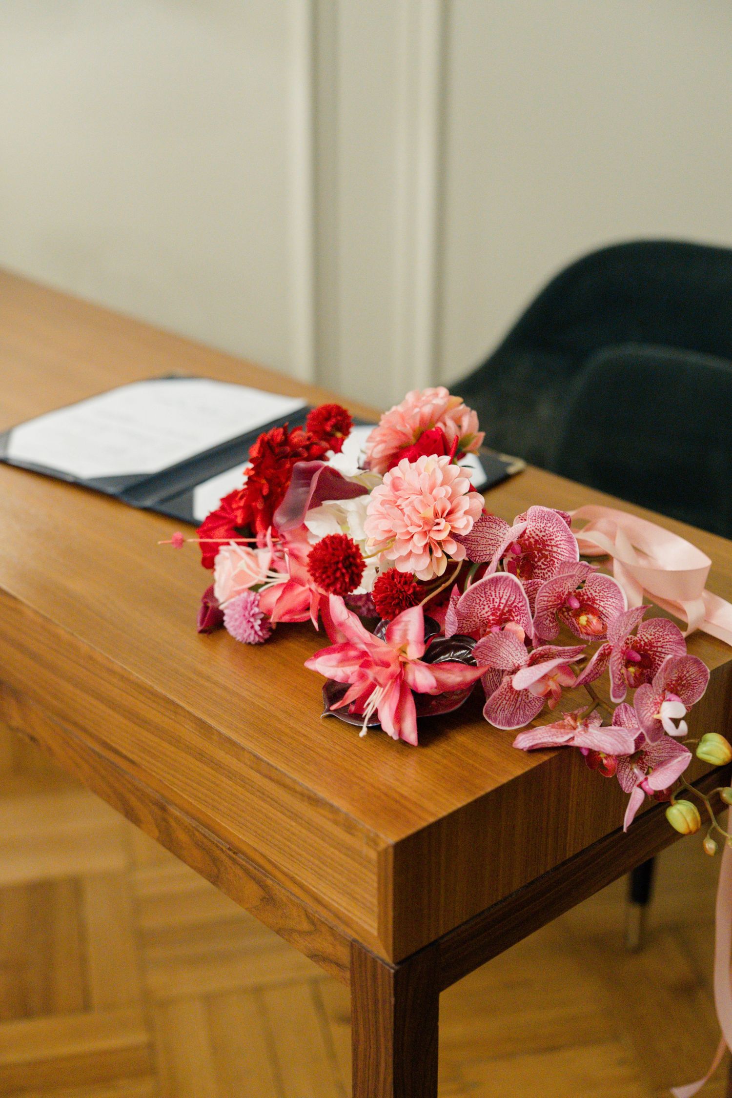 Vibrant pink and red wedding bouquet with orchids rests on wooden table beside registry documents.