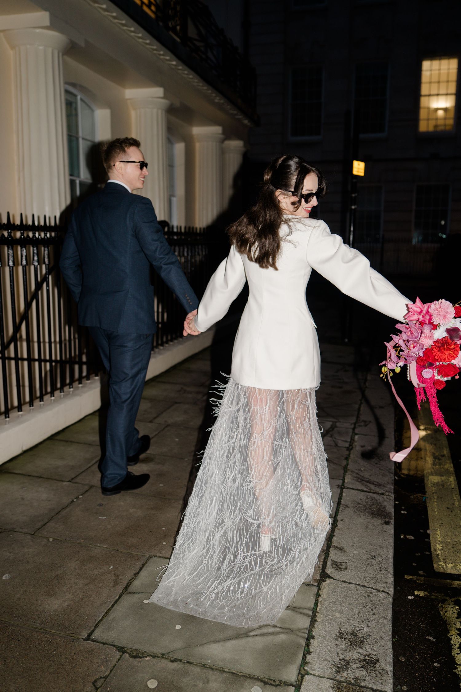 Newlyweds hold hands walking along a London street at night, bride wearing white coat and feather skirt.