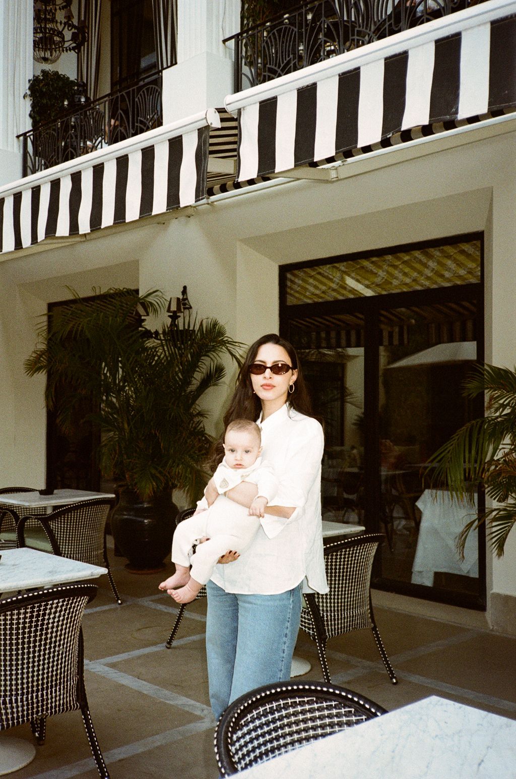 A woman in a white shirt and jeans holds a baby outside a elegant hotel patio with striped awnings.