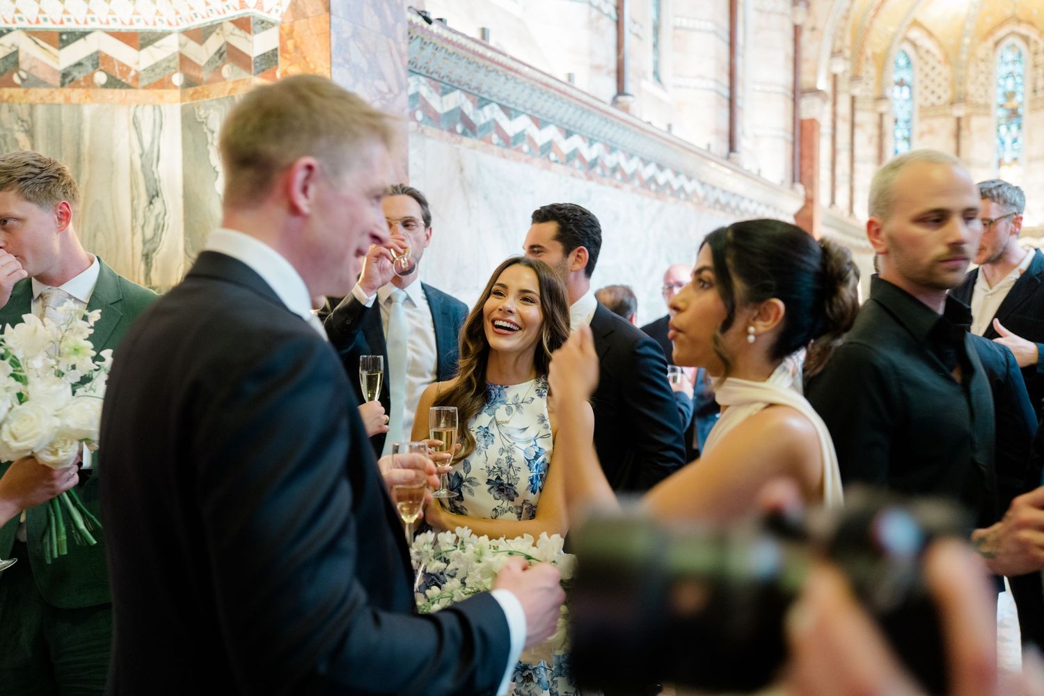 Wedding guests laughing and toasting with champagne glasses inside elegant reception venue.