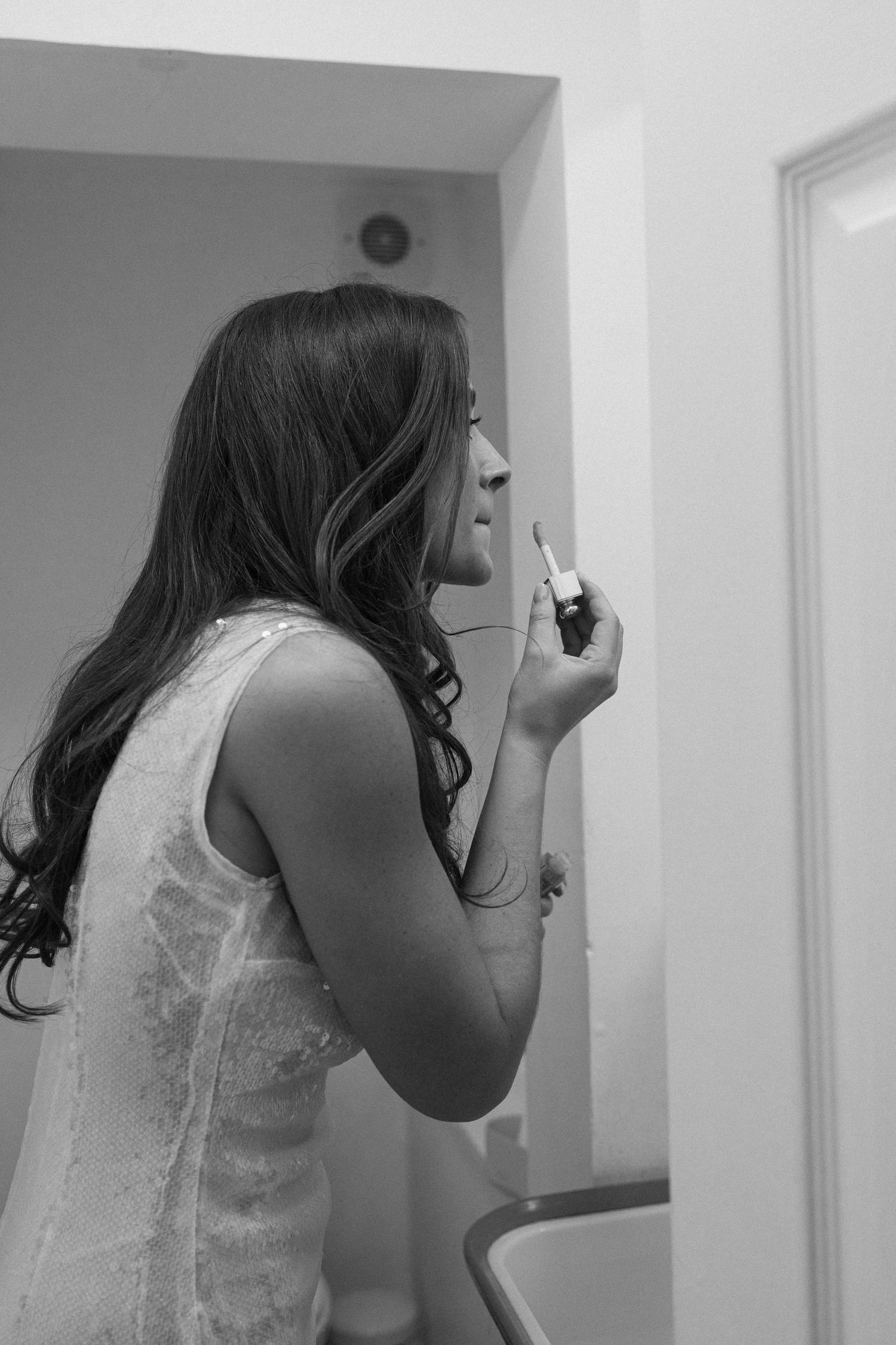 Bride in white lace dress applying lipstick in bathroom mirror while preparing for her wedding day.