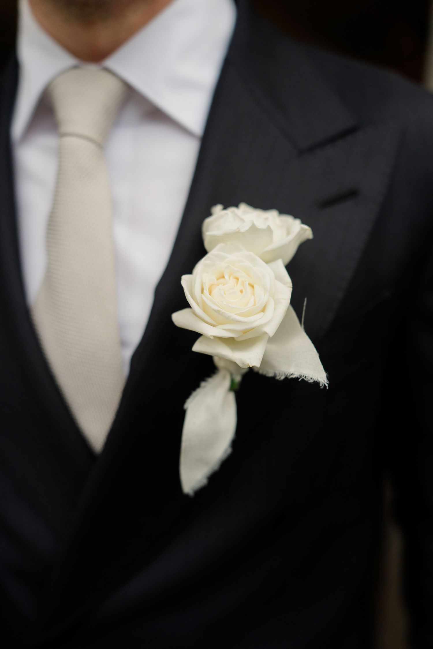 Groom wearing black suit with white rose boutonniere and cream tie at a formal wedding ceremony.