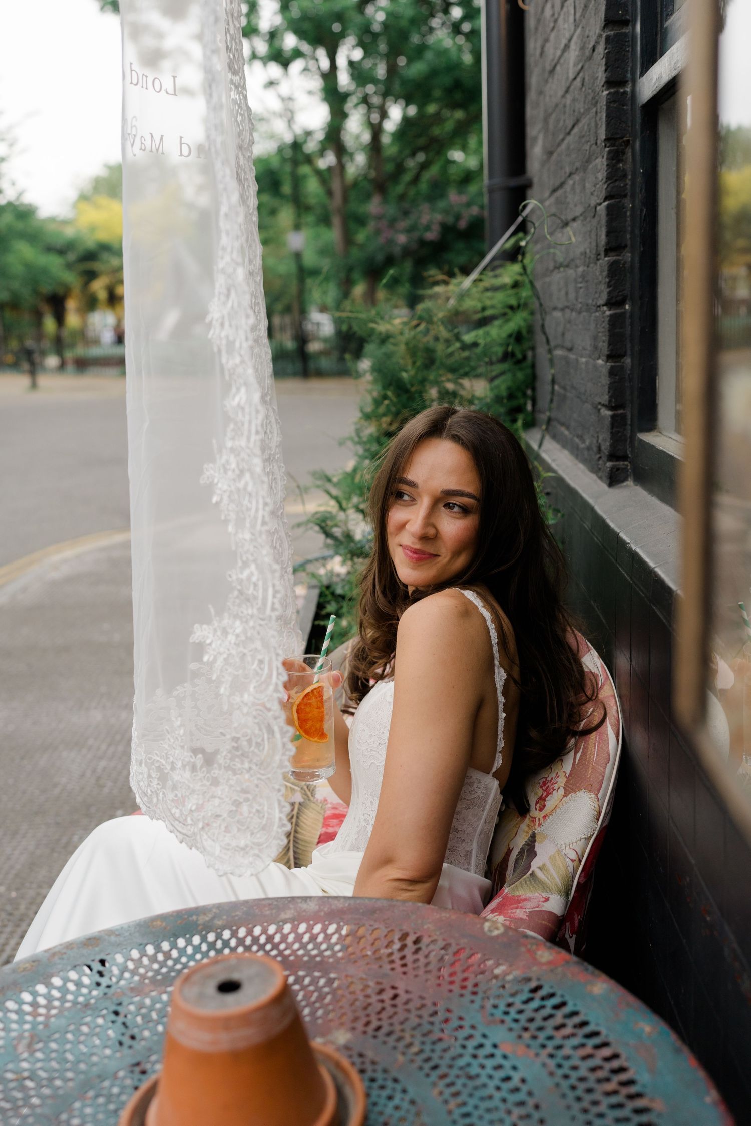 Bride in white dress sitting outdoors holding a cocktail beside a lace veil at a cafe.