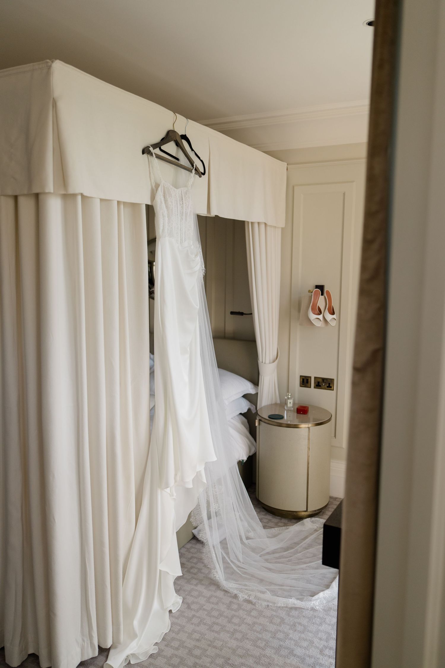 White wedding dress hanging on a four-poster bed in a luxury hotel bridal suite.