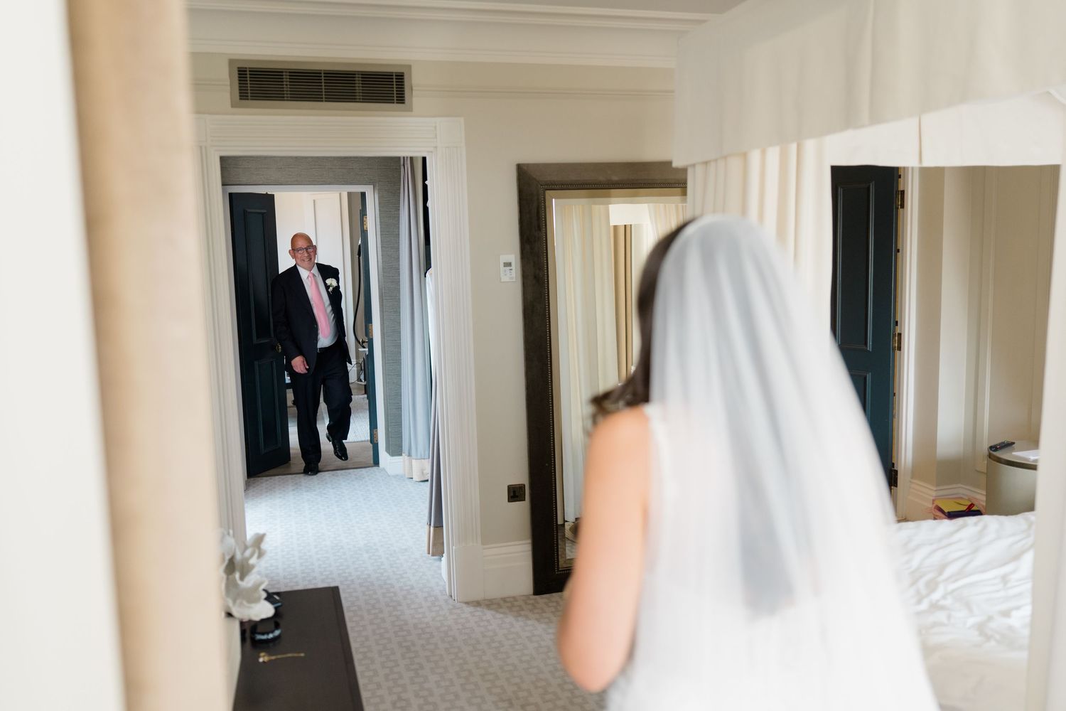 Bride in white veil stands before mirror as suited father walks through hotel room doorway.