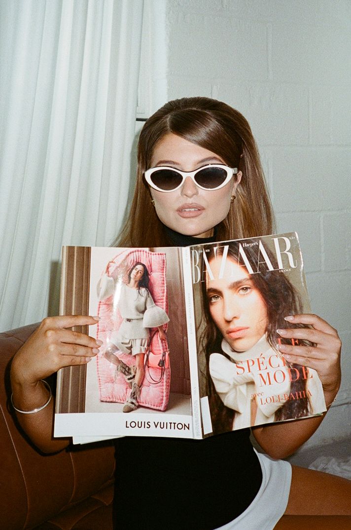 Young woman in white cat-eye sunglasses holding a Harper's Bazaar magazine open, showing a Louis Vuitton ad inside.