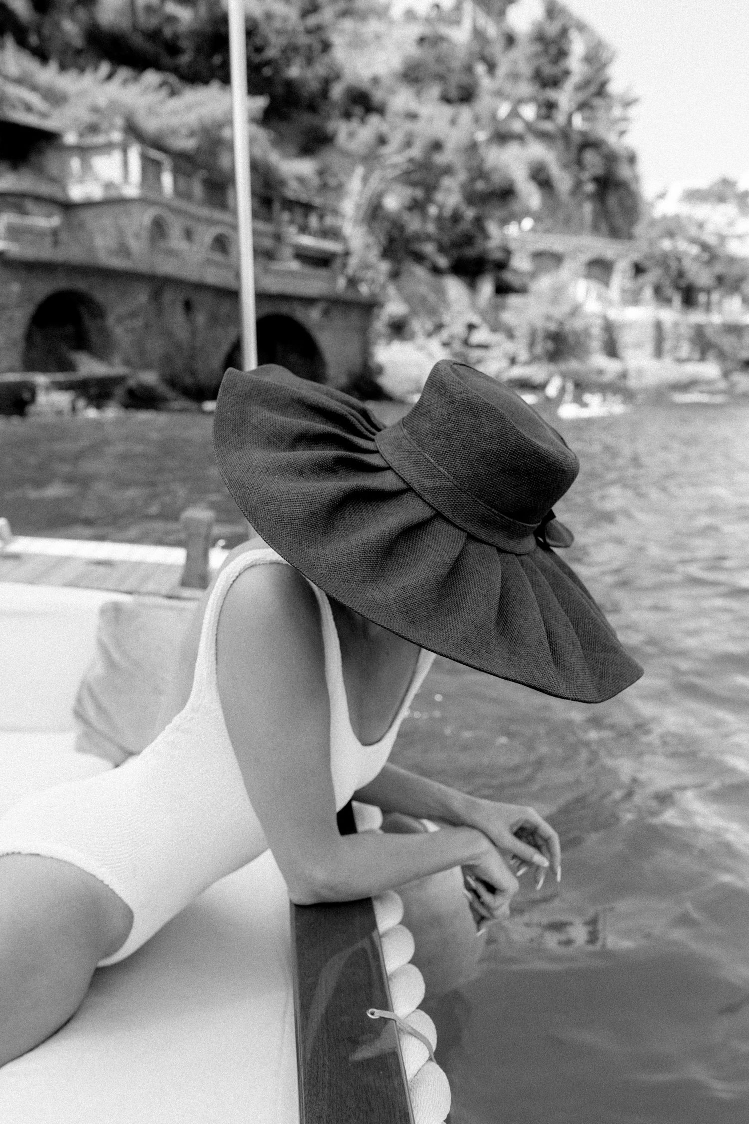Woman in white swimsuit and large black sun hat relaxing on a boat near a scenic waterfront, black and white photo.