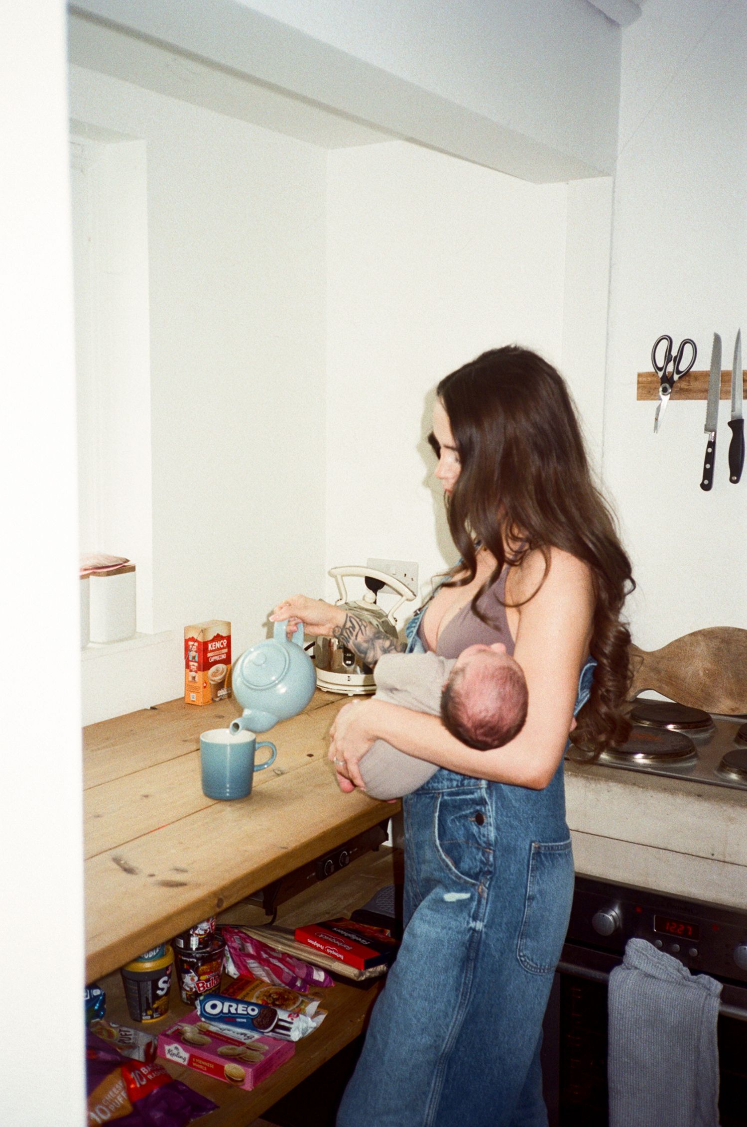 A young woman with long dark hair breastfeeds her baby while pouring tea in a cozy kitchen with Oreo cookies on the shelf.