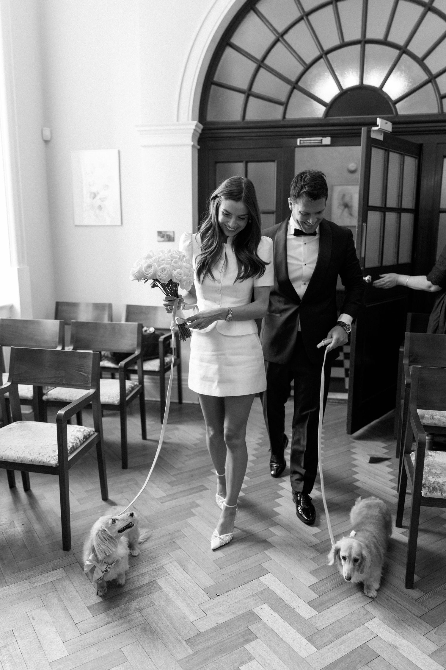 Black and white photo of bride and groom walking two dogs down the aisle in a civil ceremony venue.