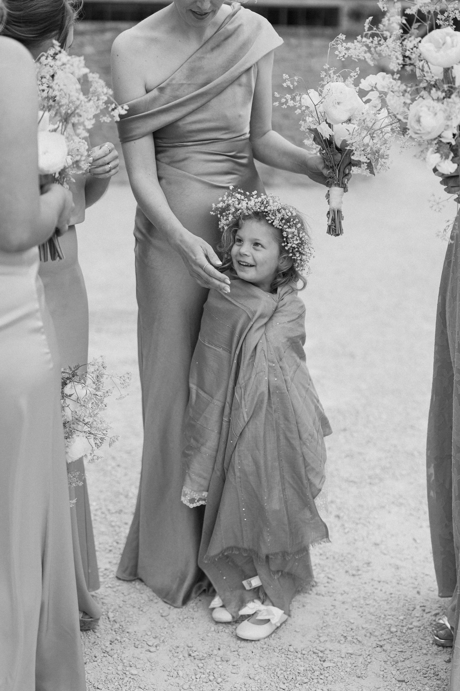 Adorable flower girl in floral crown hugs bridesmaid's leg at outdoor wedding ceremony.