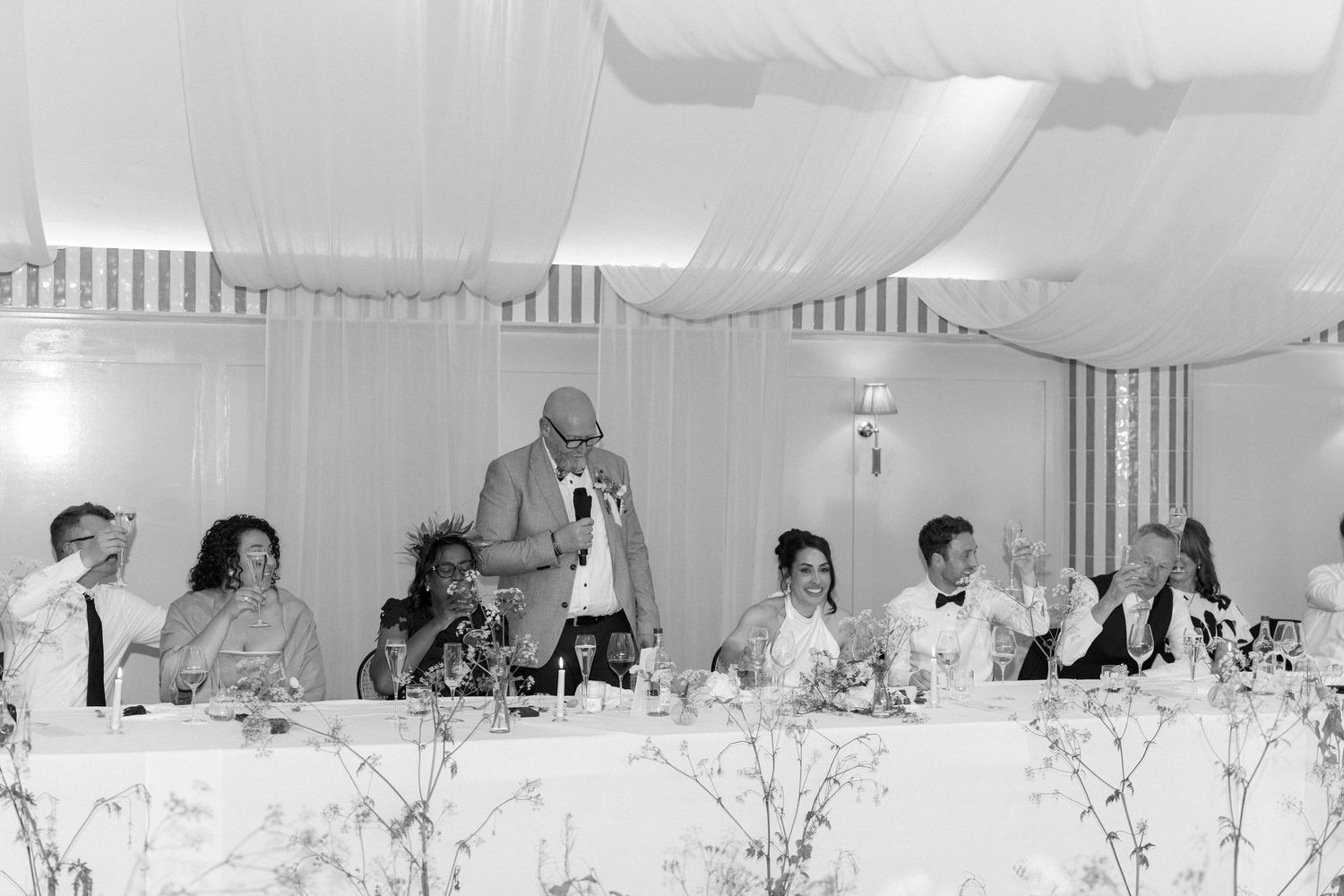 Wedding party seated at head table as guest delivers speech in elegantly draped reception hall.