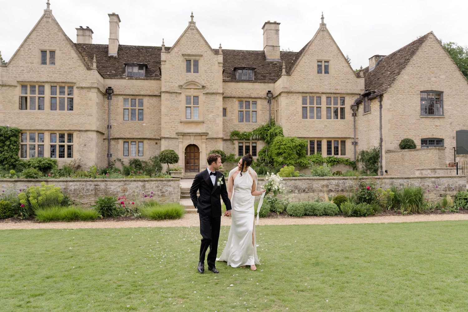 Bride and groom walk hand in hand across lawn in front of picturesque Cotswolds stone country house venue.