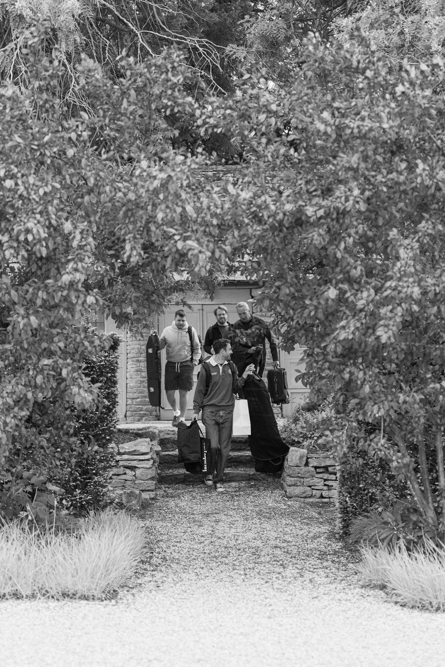 Black and white photo of a wedding party walking through a tree-lined stone archway entrance carrying luggage.