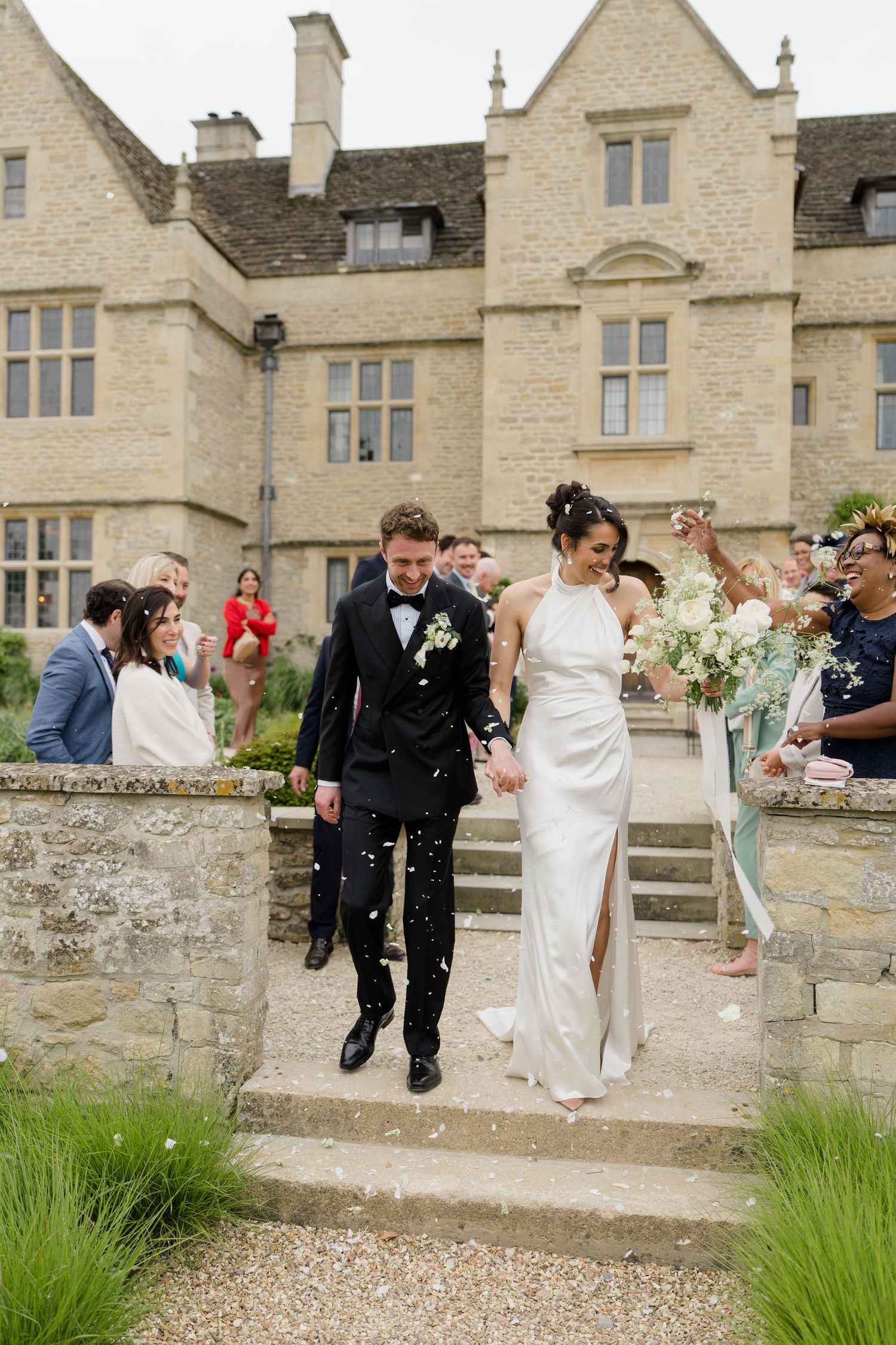 Bride and groom walk down stone steps as guests throw confetti outside a stunning Cotswolds manor wedding venue.