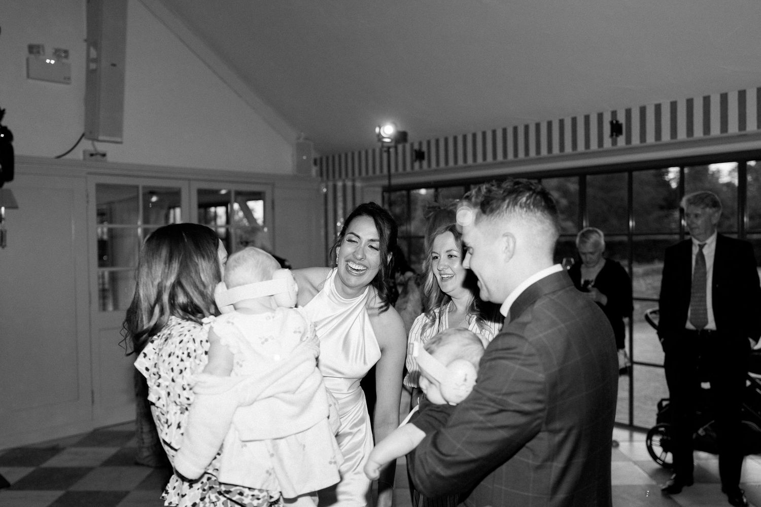 Bride smiles joyfully while guests admire babies at indoor wedding reception in black and white photo.