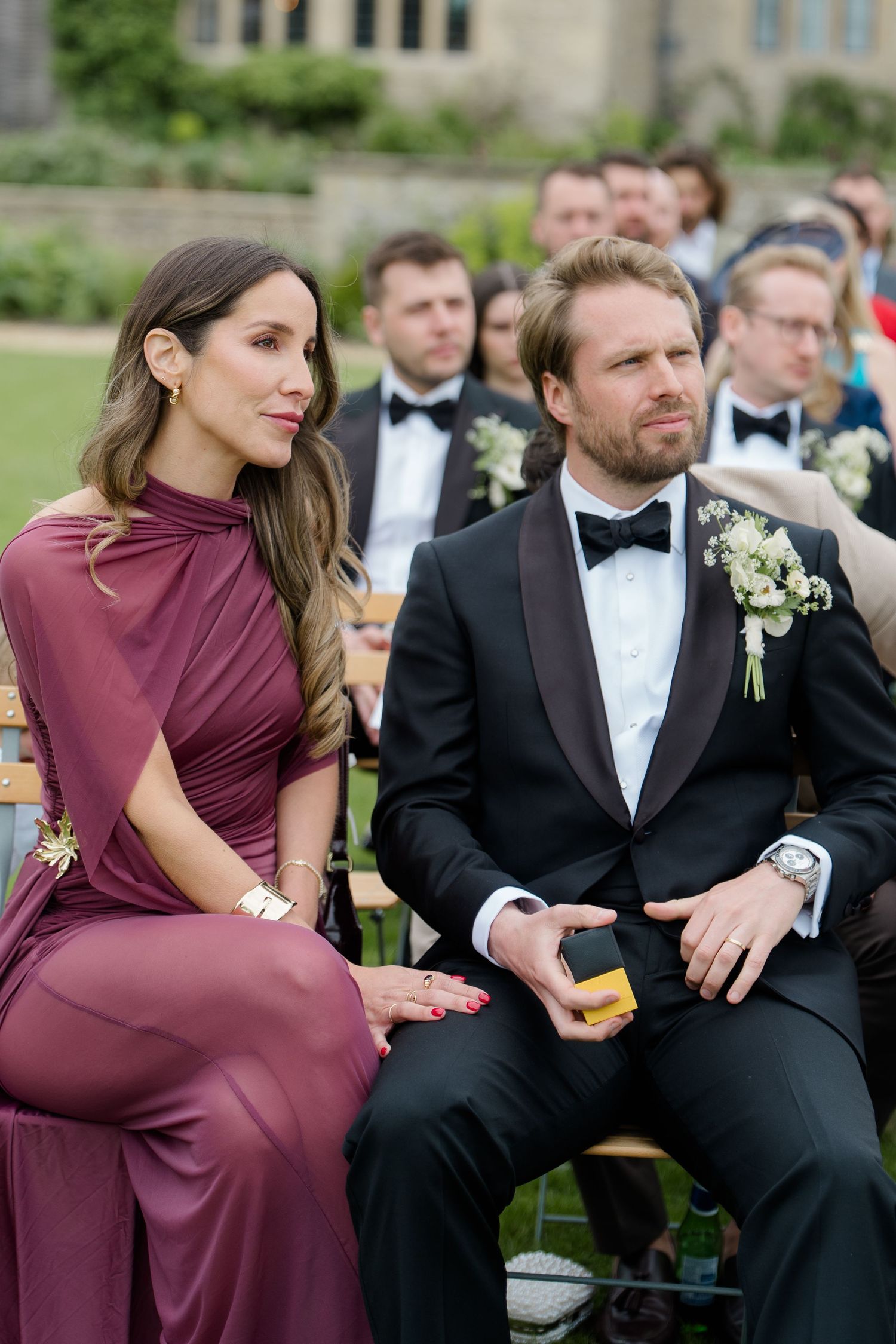 Elegant wedding guest in burgundy gown seated beside groomsman with boutonniere at ceremony.