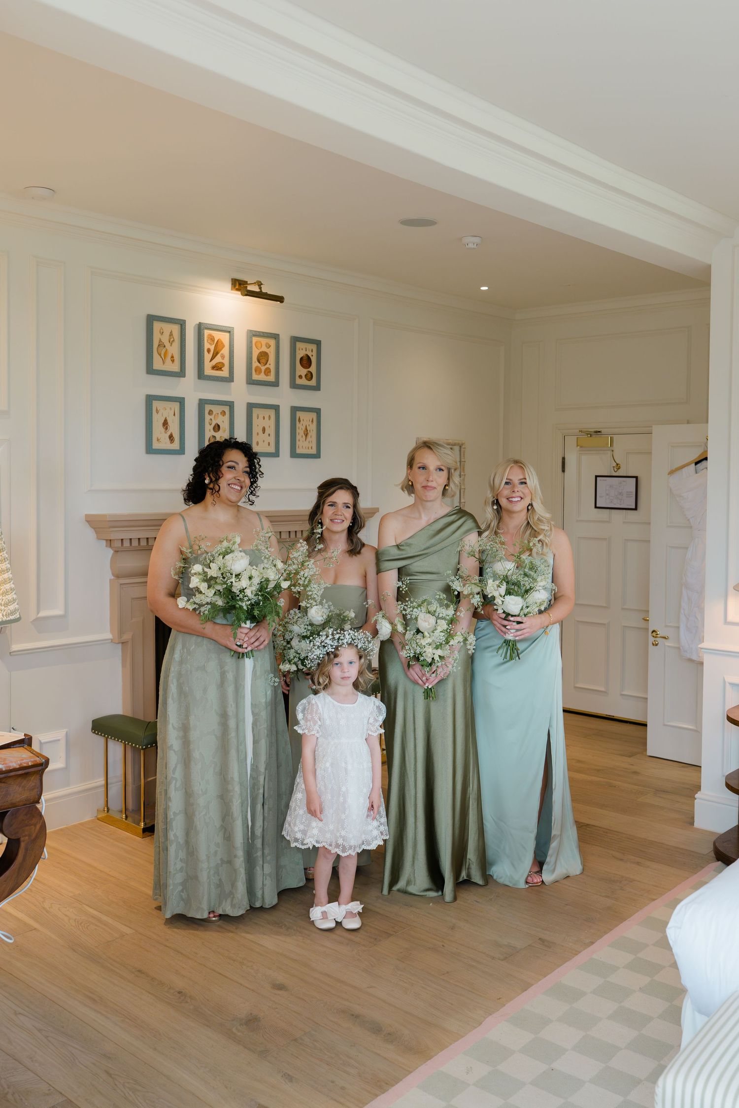 Four bridesmaids in sage green dresses with a flower girl pose indoors holding white floral bouquets.