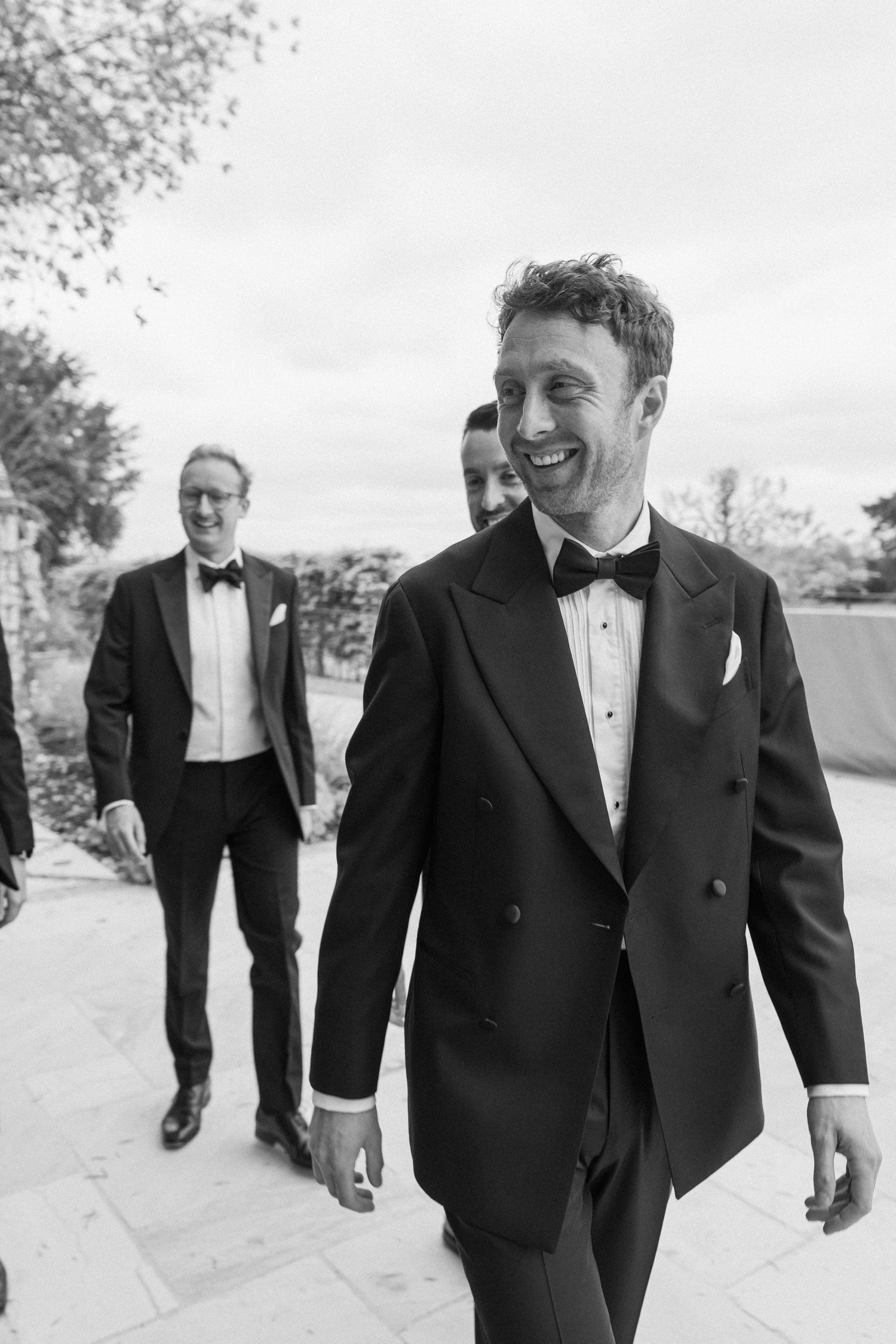 Black and white photo of smiling groom in double-breasted tuxedo walking with groomsmen on a stone terrace.