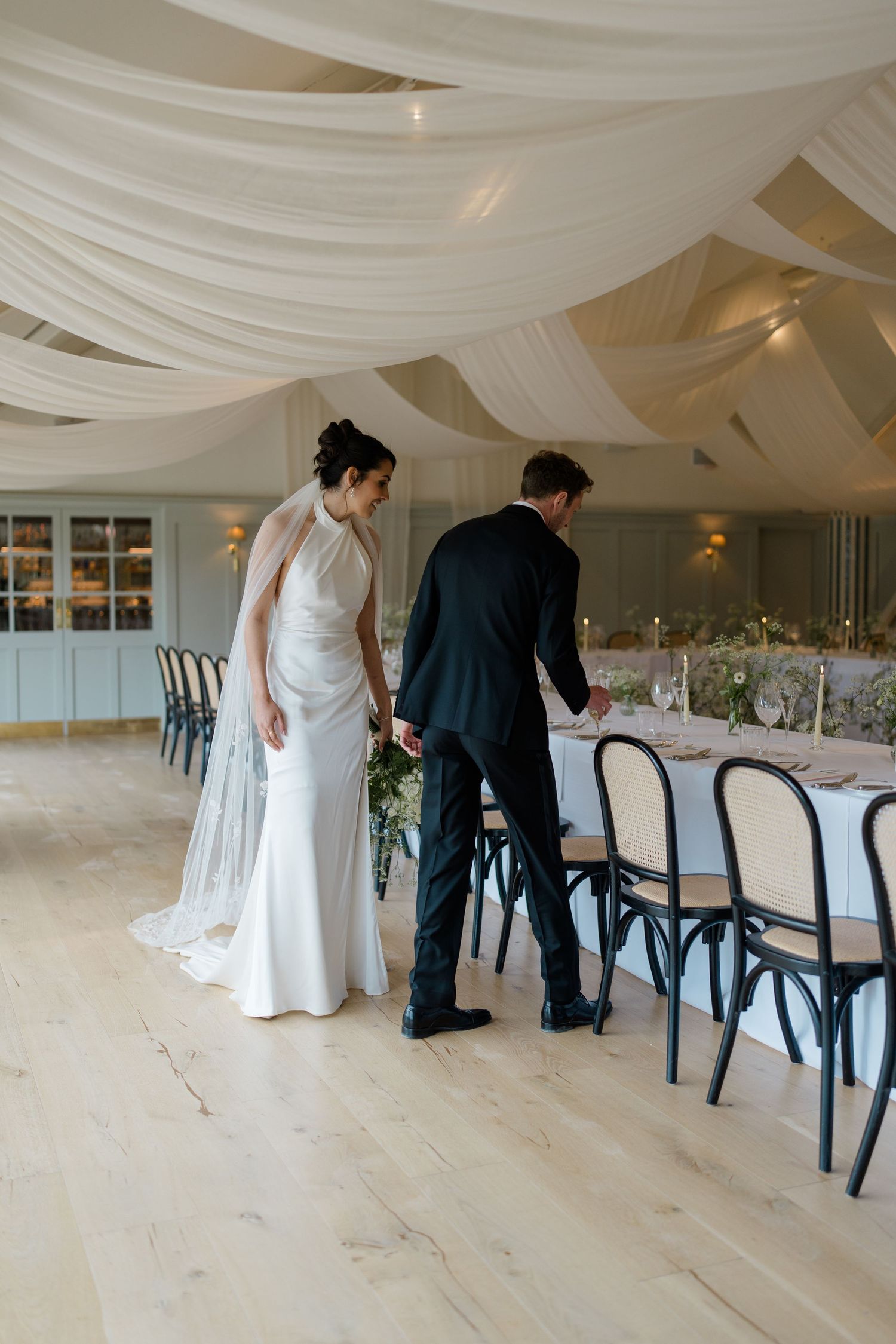 Bride and groom in white draped wedding reception venue inspecting long elegantly set dining table.