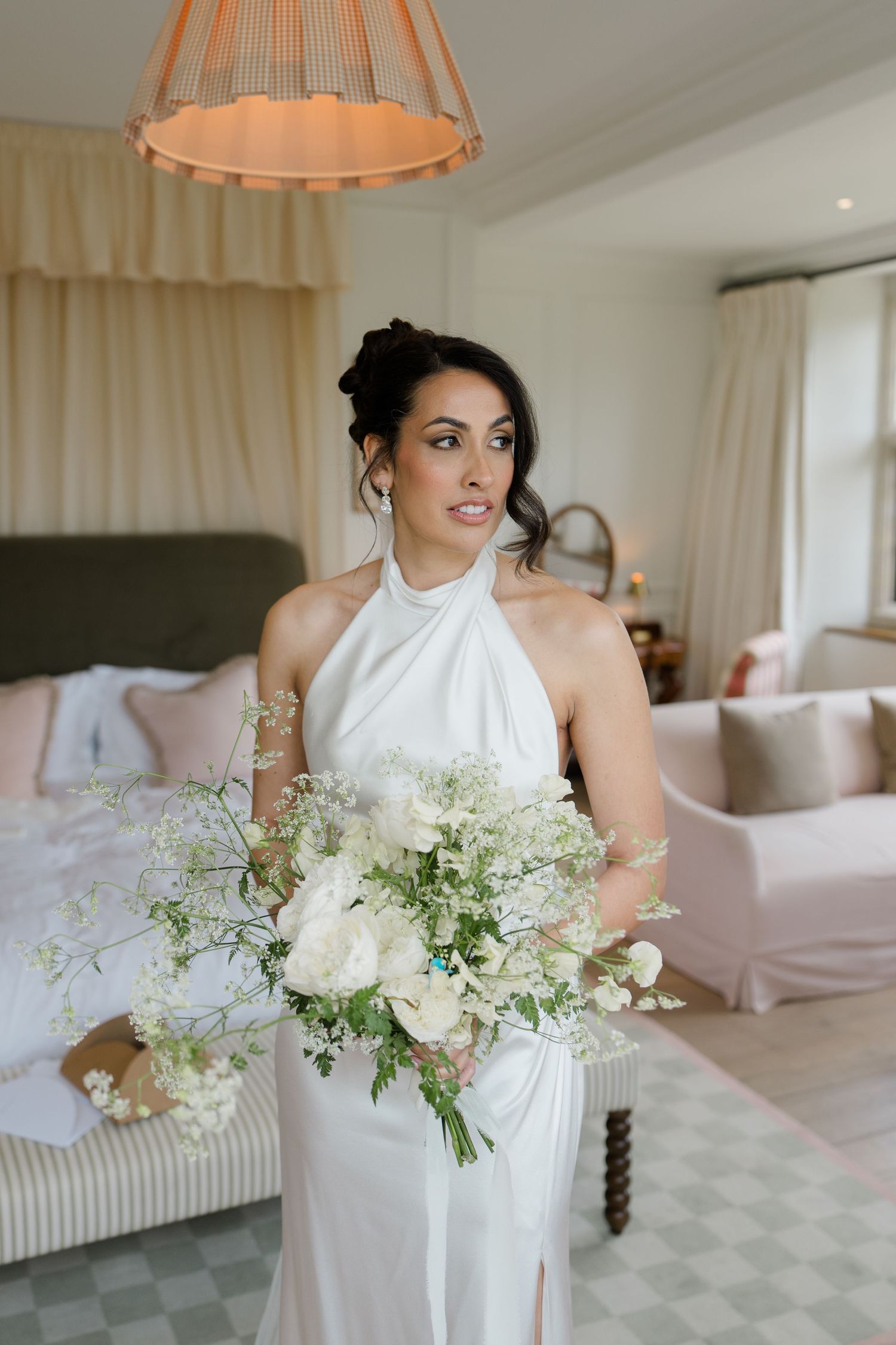Elegant bride in halter neck white gown holds large white floral bouquet in a beautifully decorated room.