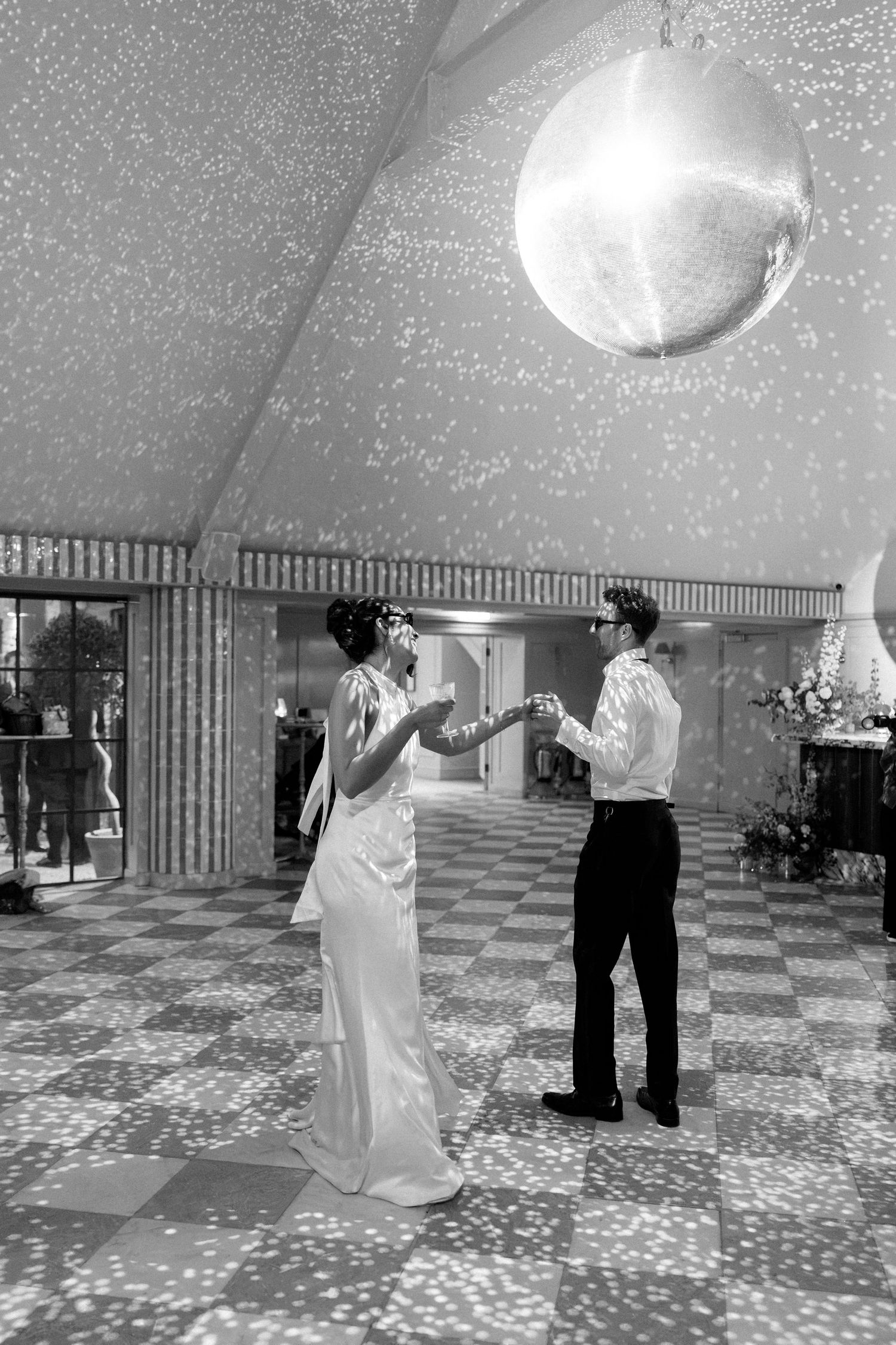 Bride and groom share first dance under sparkling disco ball on checkered floor at wedding reception.