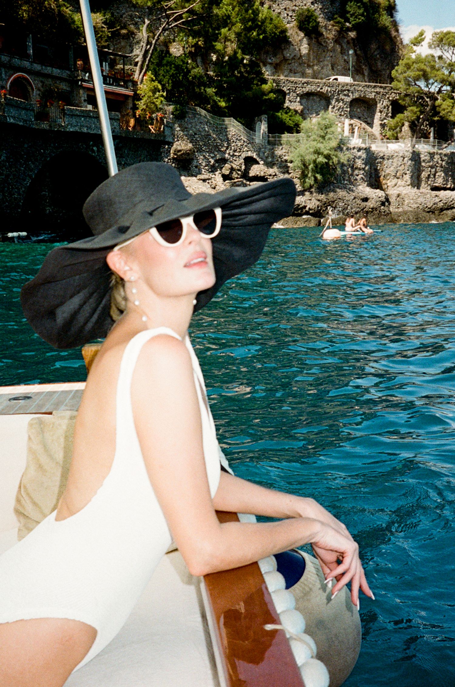 Woman in white swimsuit and black sun hat on a boat with turquoise Mediterranean waters and rocky cliffs behind her.