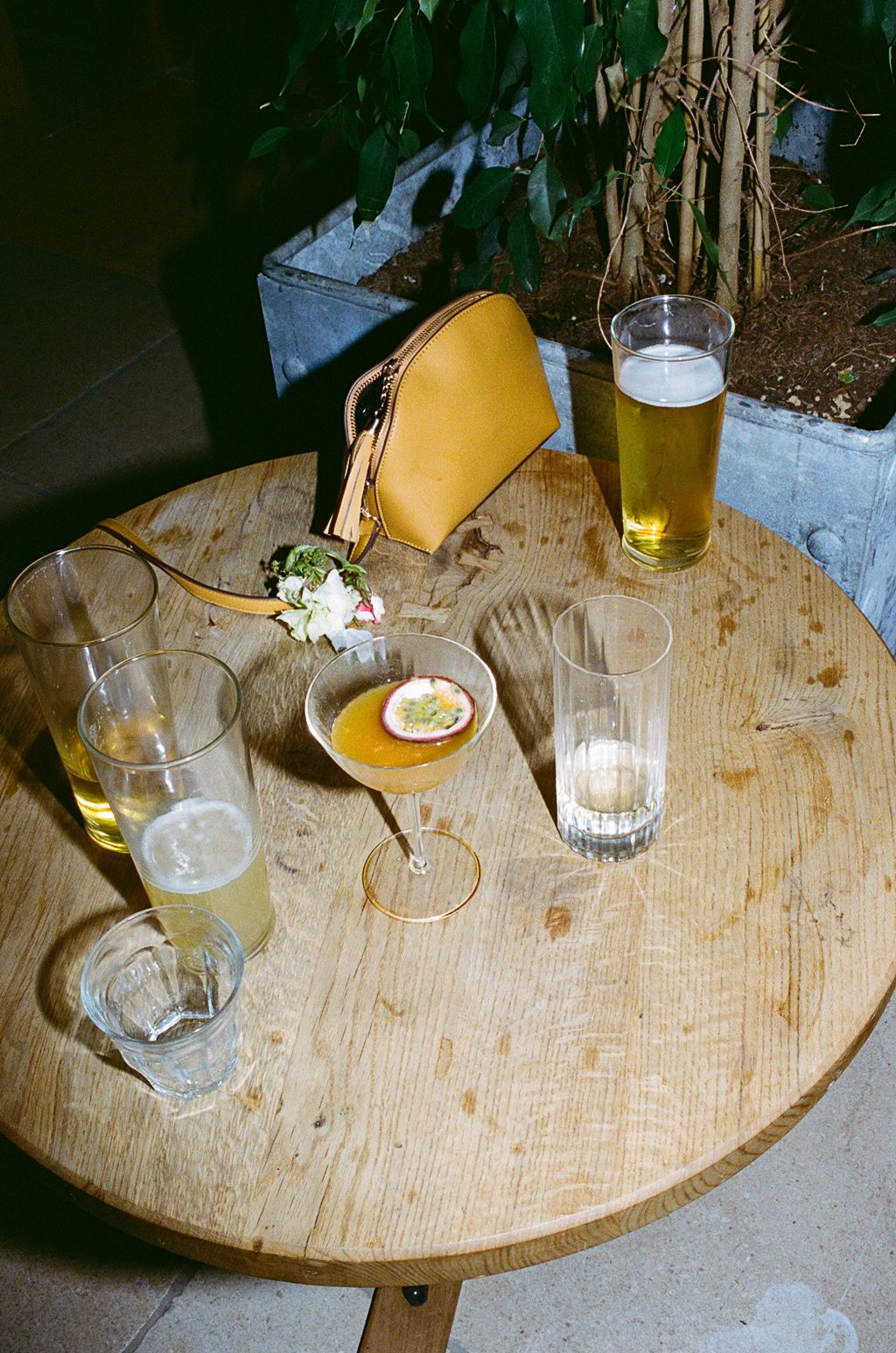 Round wooden table with cocktails, beer, white wine, and a yellow handbag at an outdoor bar setting.