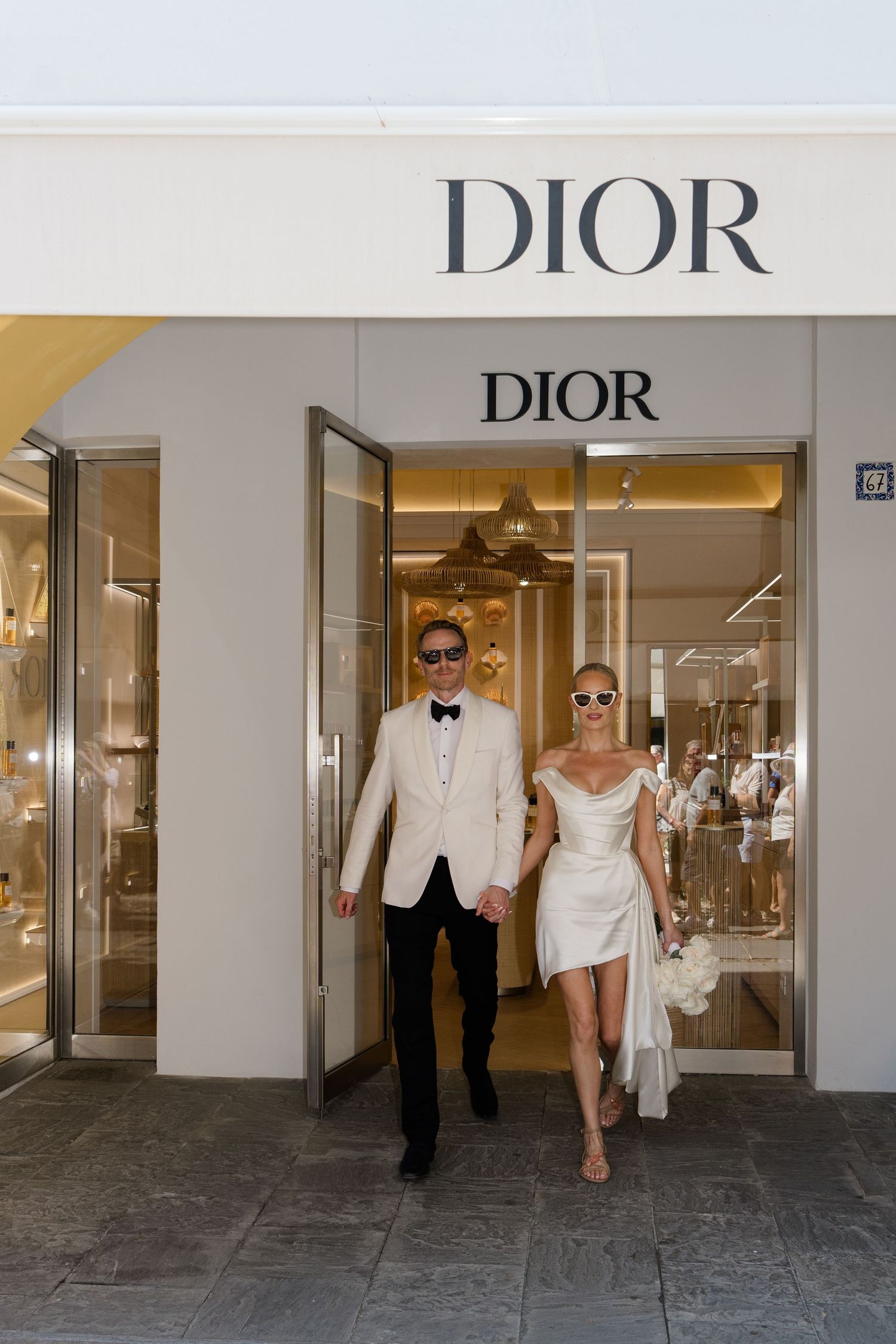 Bride and groom in white attire holding hands while exiting a Dior luxury boutique store.
