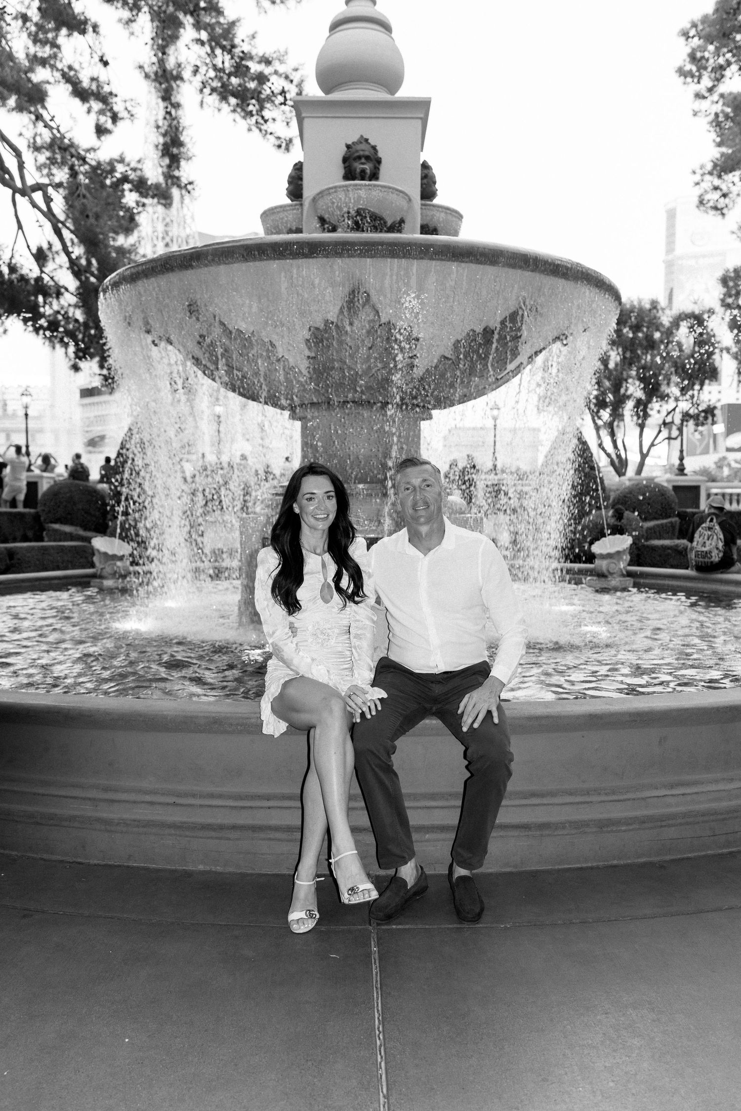 A couple poses together on the edge of an outdoor fountain.