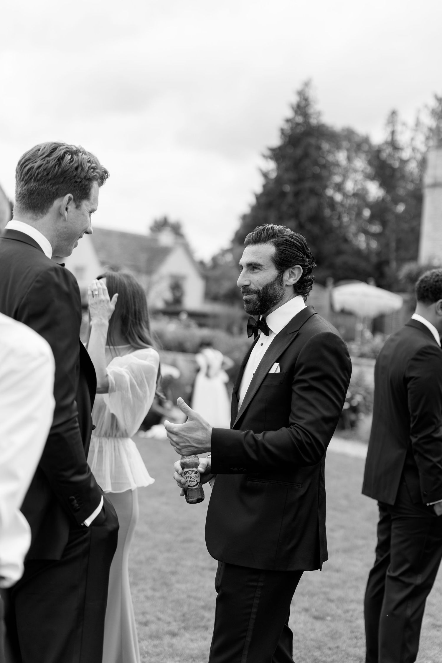 Black and white photo of elegantly dressed wedding guests conversing outdoors at a garden reception.