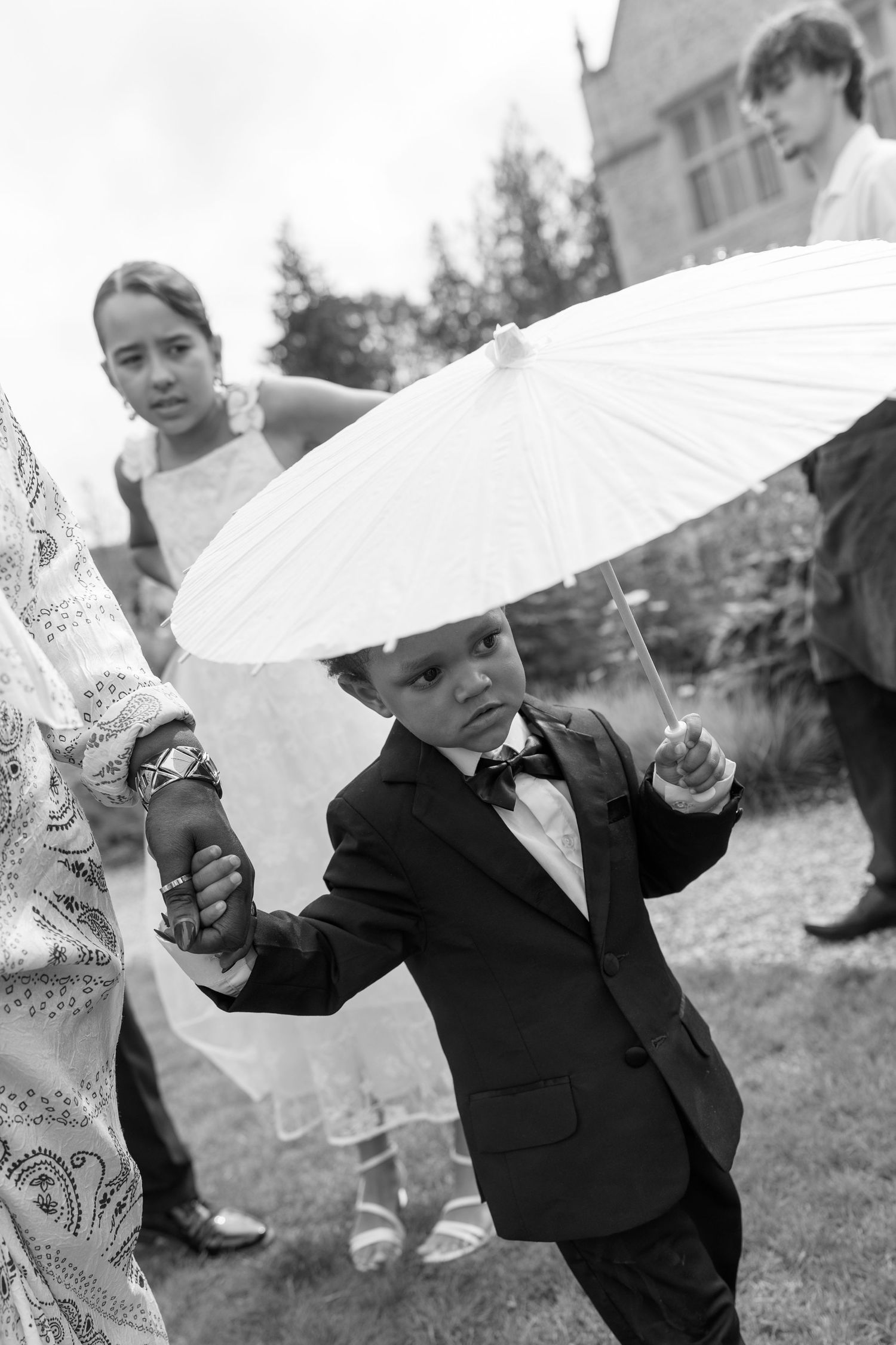 Adorable young ring bearer in tuxedo holds white parasol at outdoor wedding ceremony.