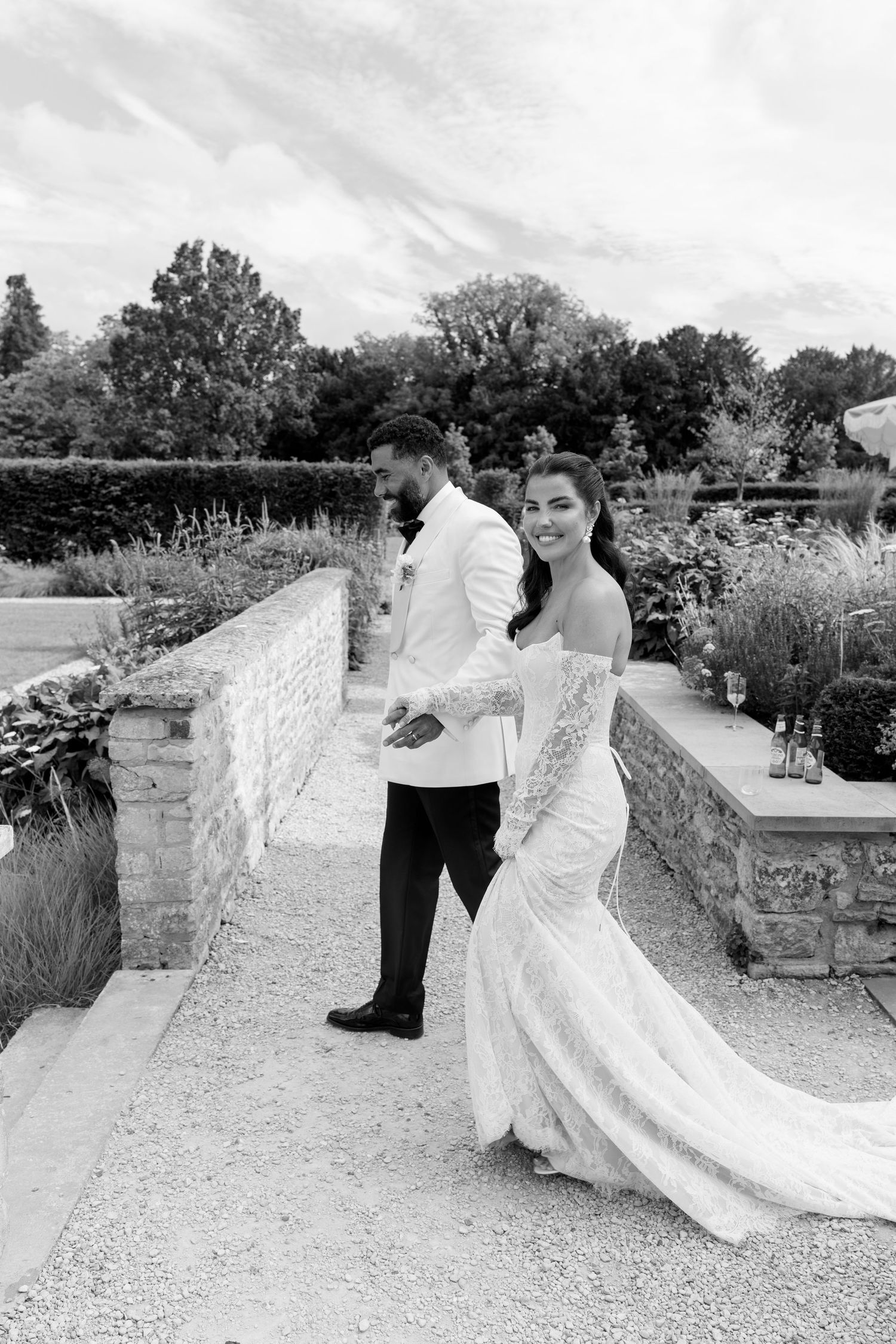 Black and white portrait of a smiling bride in lace gown and groom in white jacket strolling through a walled garden.