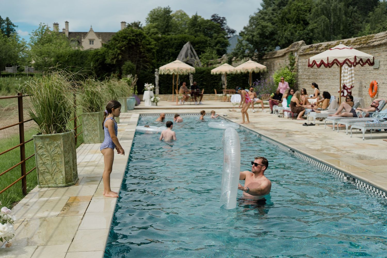 Wedding guests enjoy outdoor swimming pool with fountain at rustic countryside venue on sunny day.