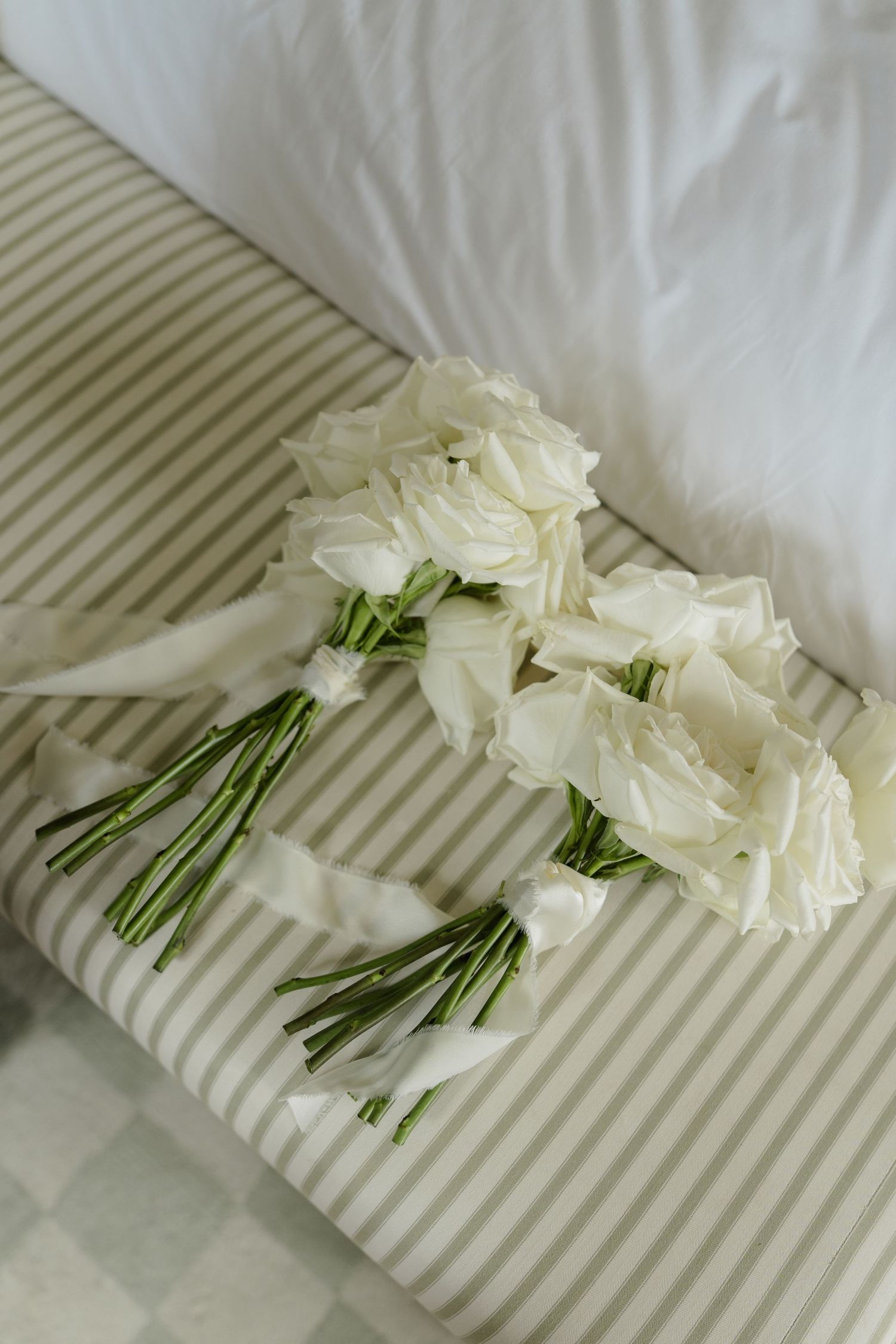 Two elegant white rose bridal bouquets with satin ribbons resting on a striped bedspread.