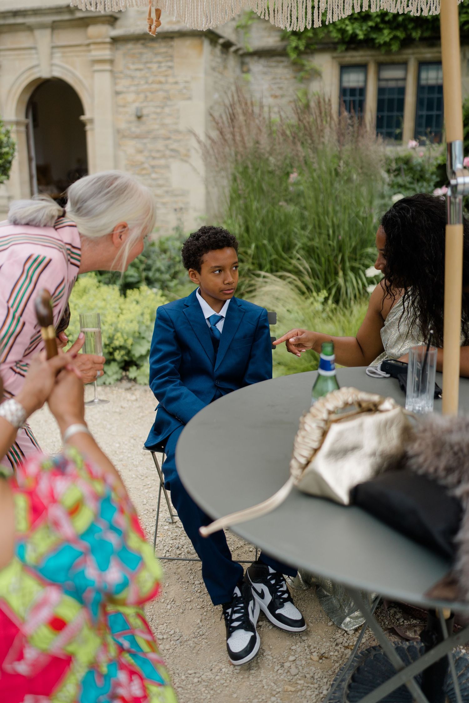 A young boy in a blue suit sits at an outdoor table during a wedding reception, engaged in conversation with guests.