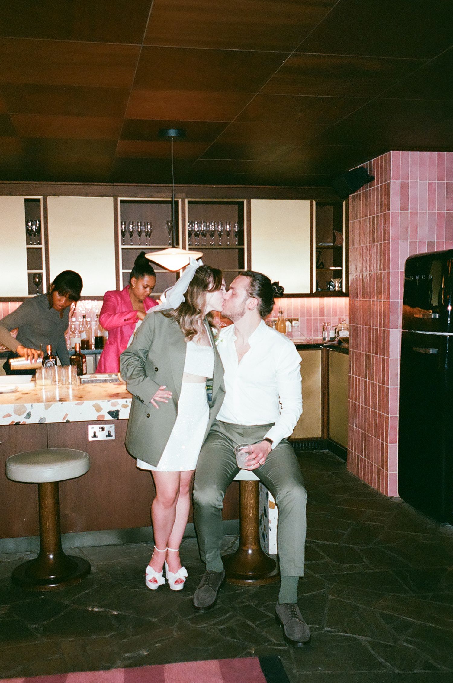 Retro-style photo of people socializing at a bar with pink tile walls and dark wood ceiling.