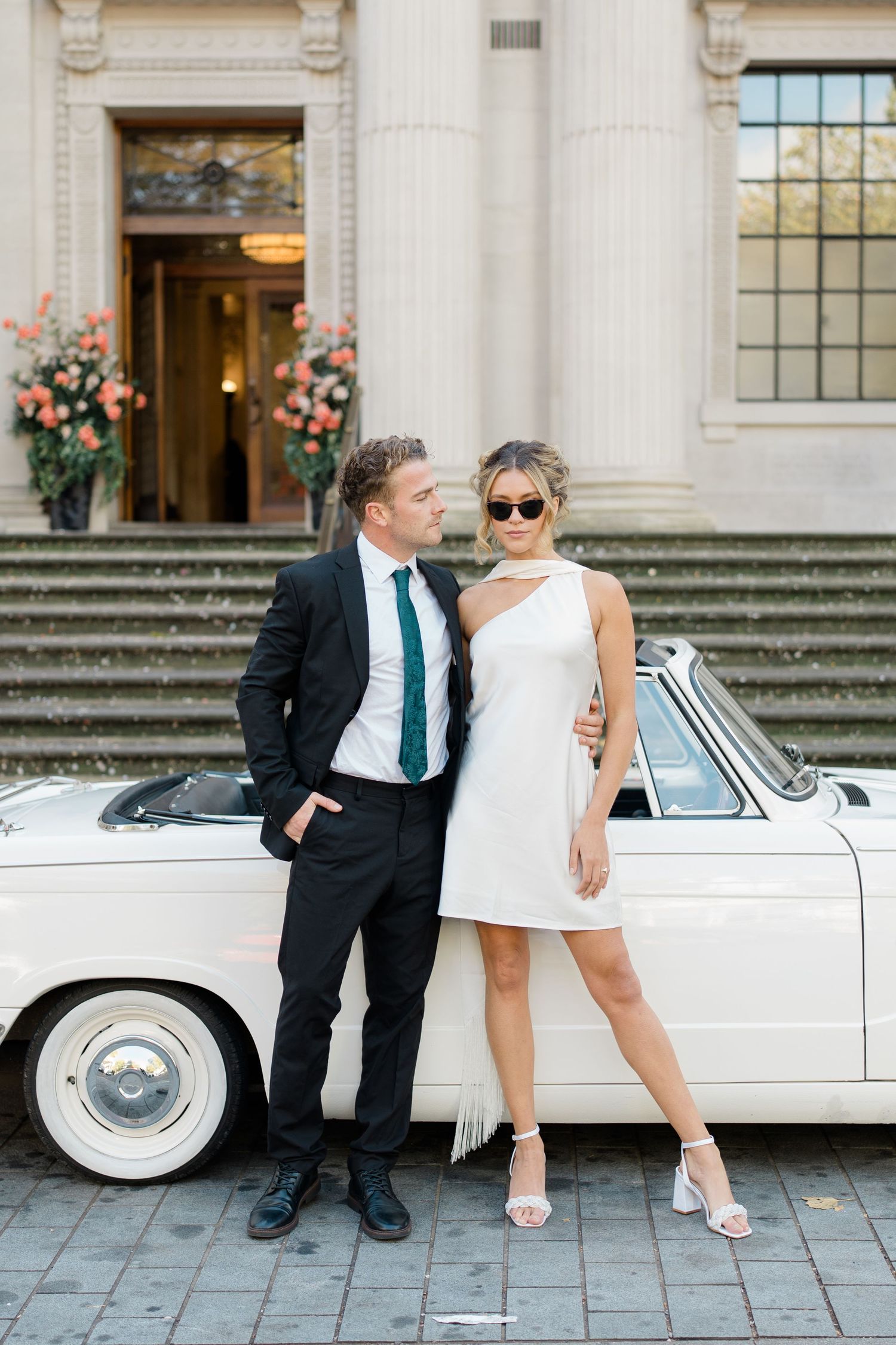 Stylish couple poses beside classic white car on stone steps of elegant building.