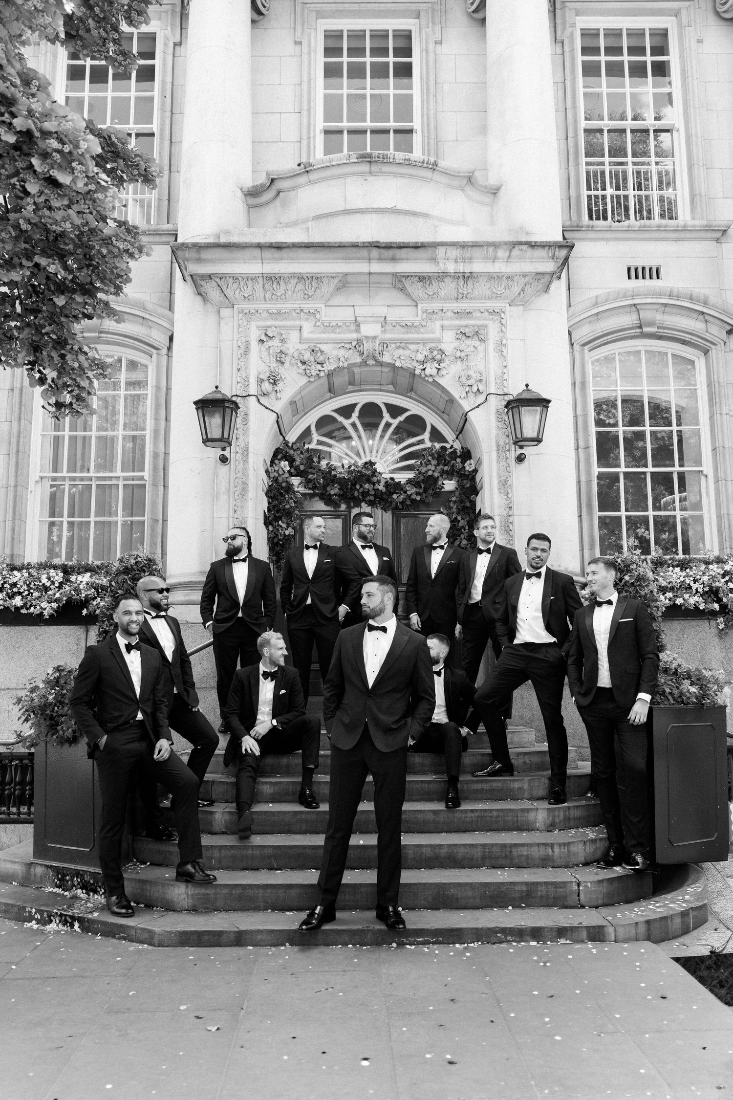 Groomsmen in black tuxedos pose on grand stone steps of historic building.