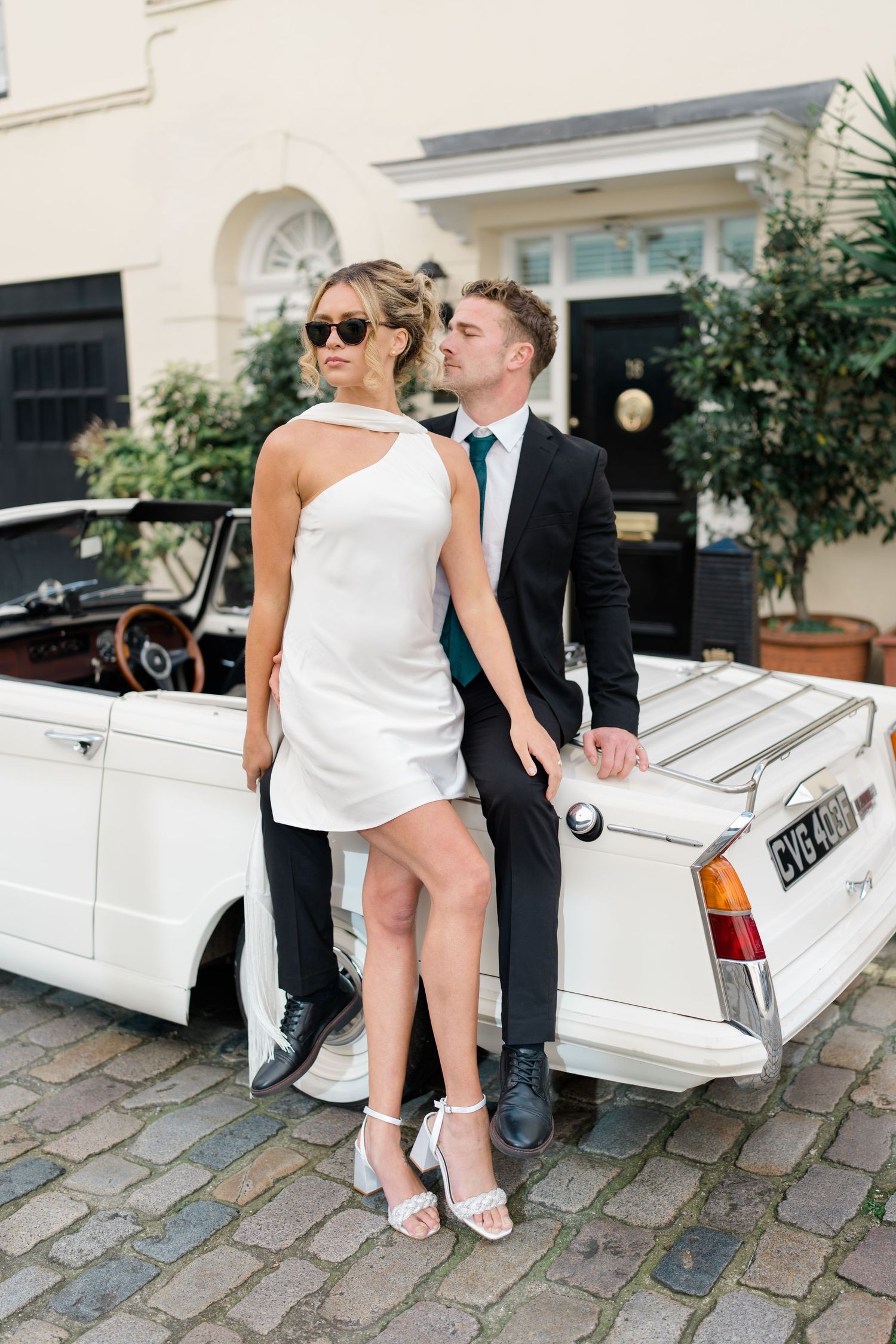 A couple sits on the back of a white vintage car on a cobblestone street.