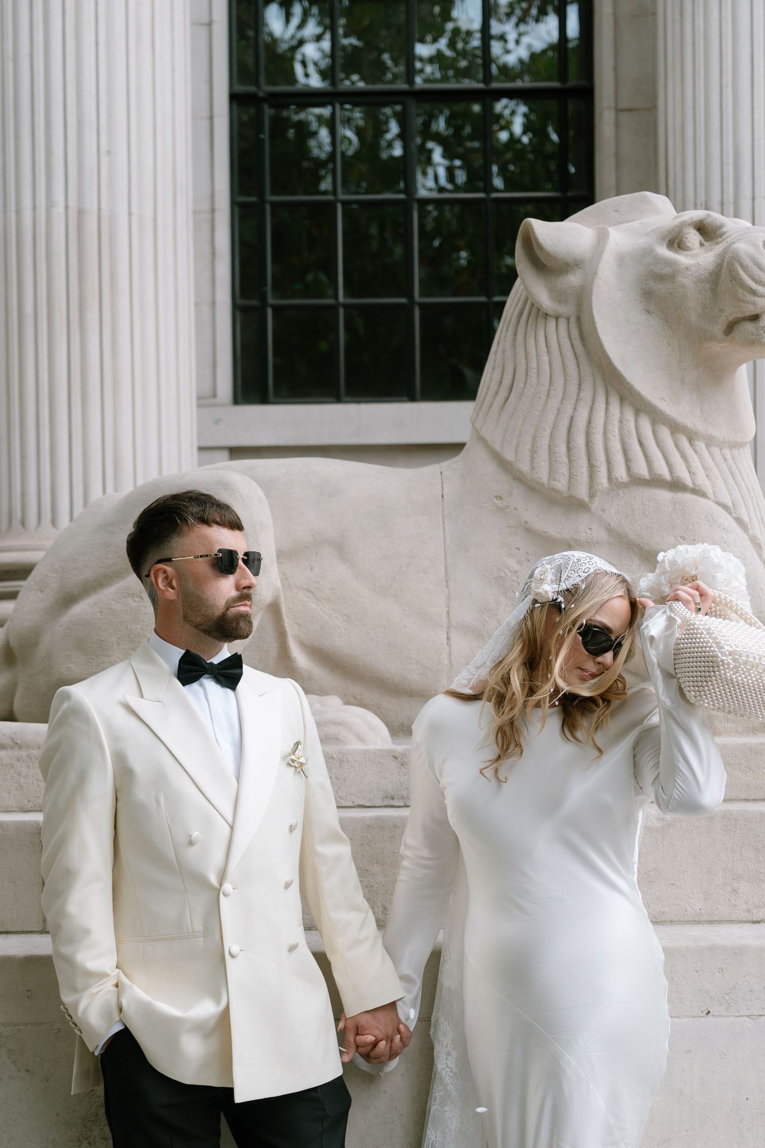 Couple in white formal attire poses against classical stone lion sculpture.