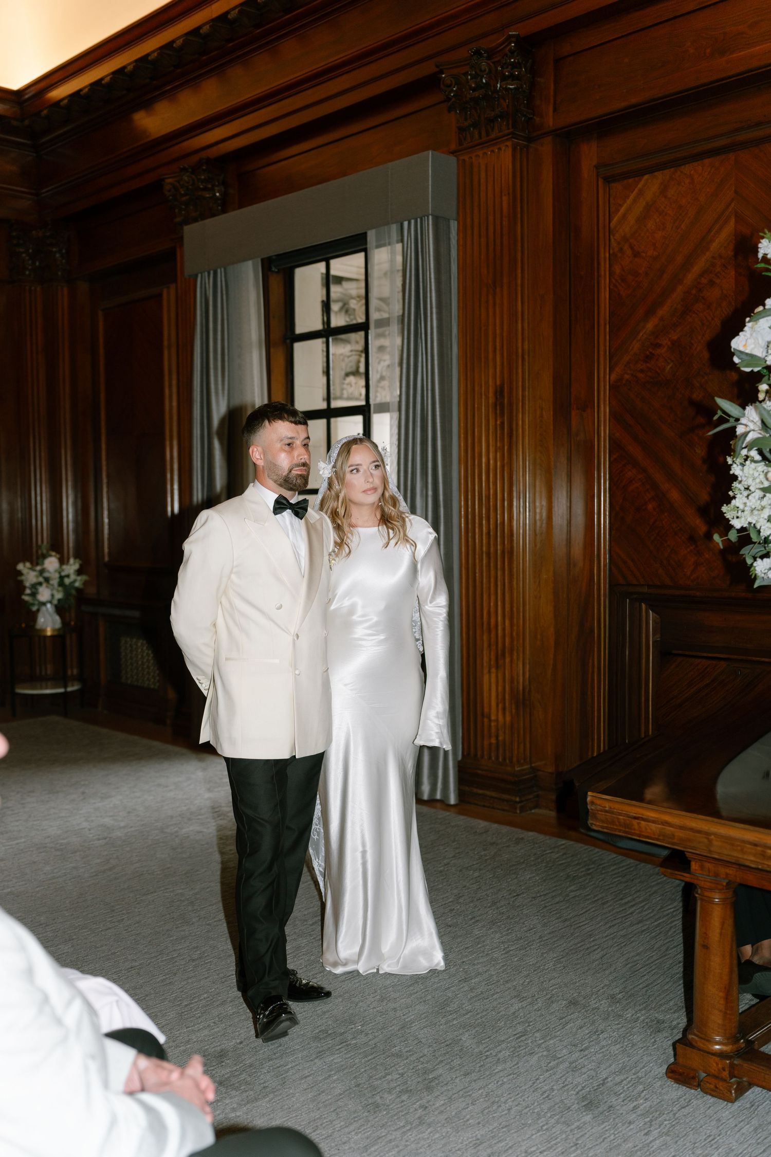 A couple in wedding attire dances in a classic wood-paneled room with large windows.