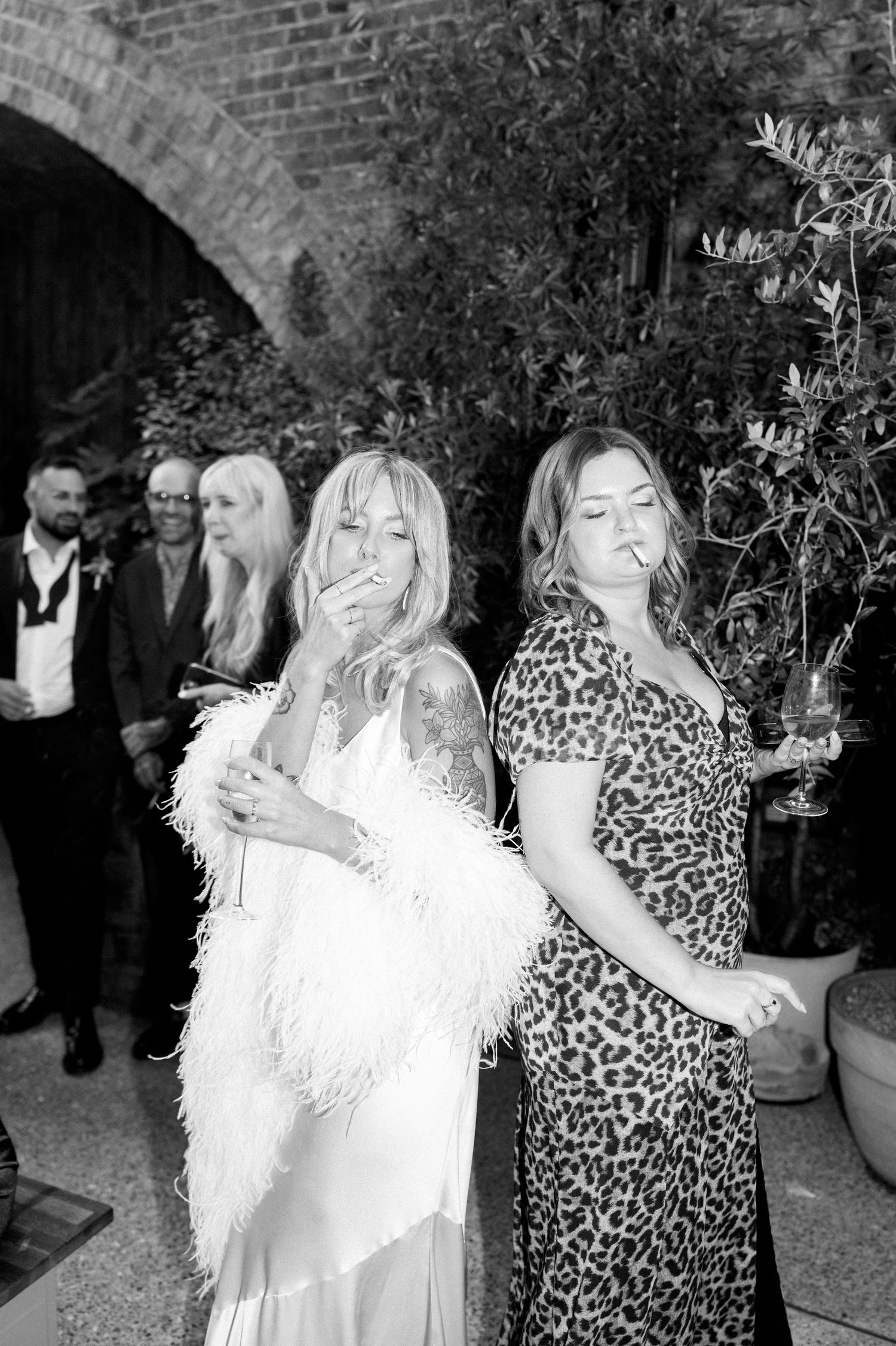 Party guests in formal attire and feather boas enjoying drinks and conversation against a brick wall backdrop.
