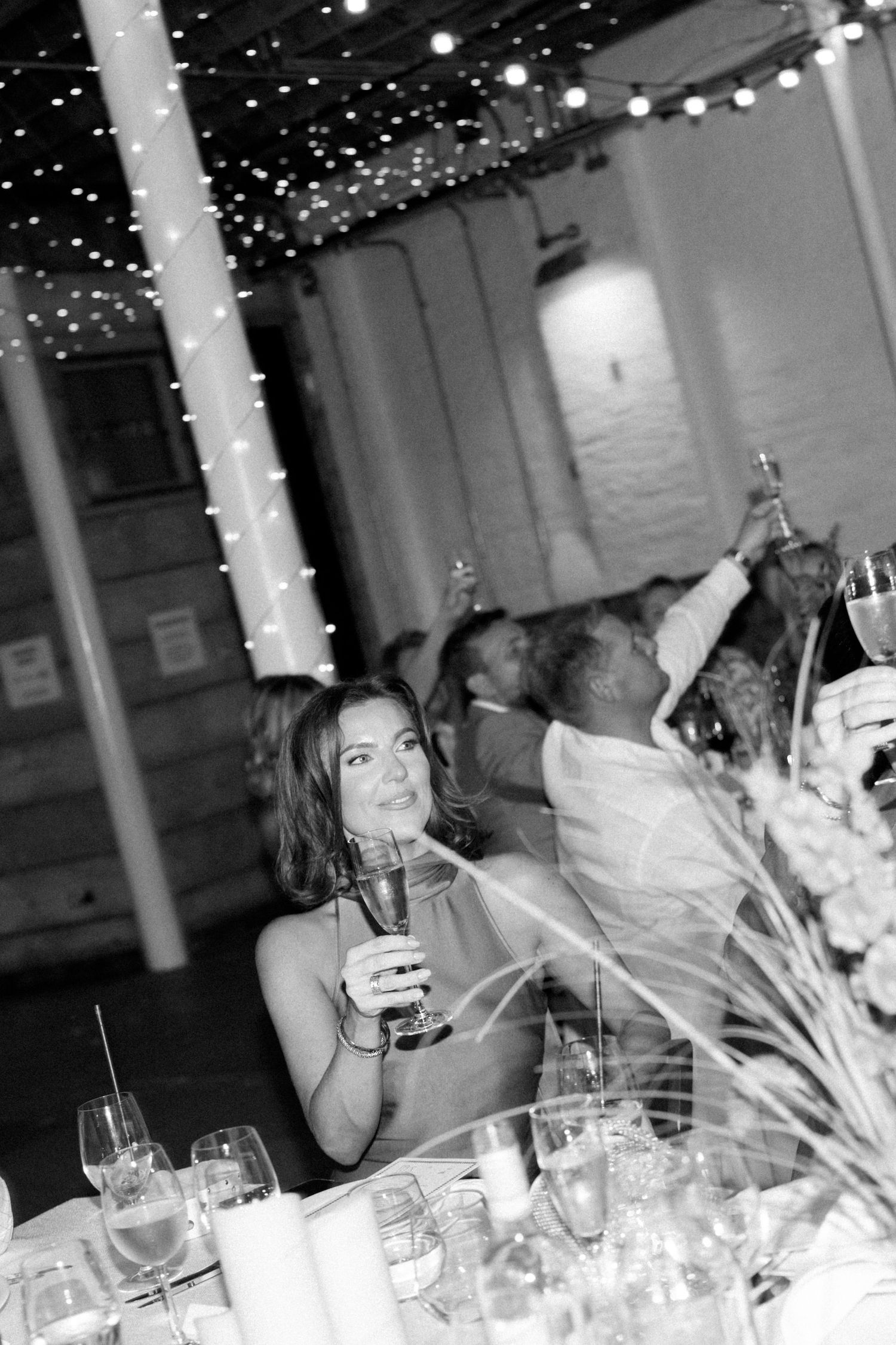 Wedding reception under twinkling lights with guests raising glasses in celebration.