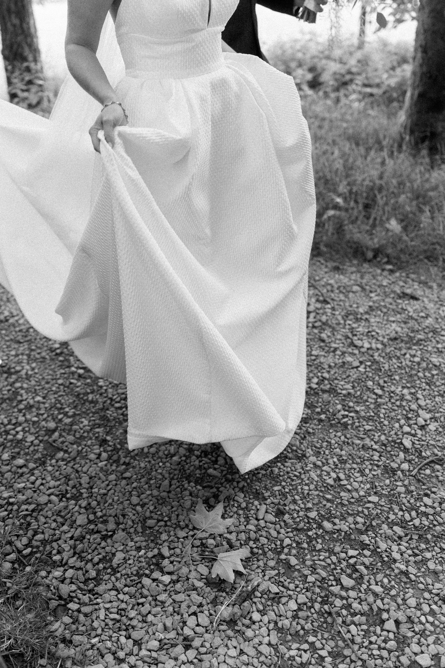 Close-up black and white shot of a flowing white wedding dress skirt on gravel ground.
