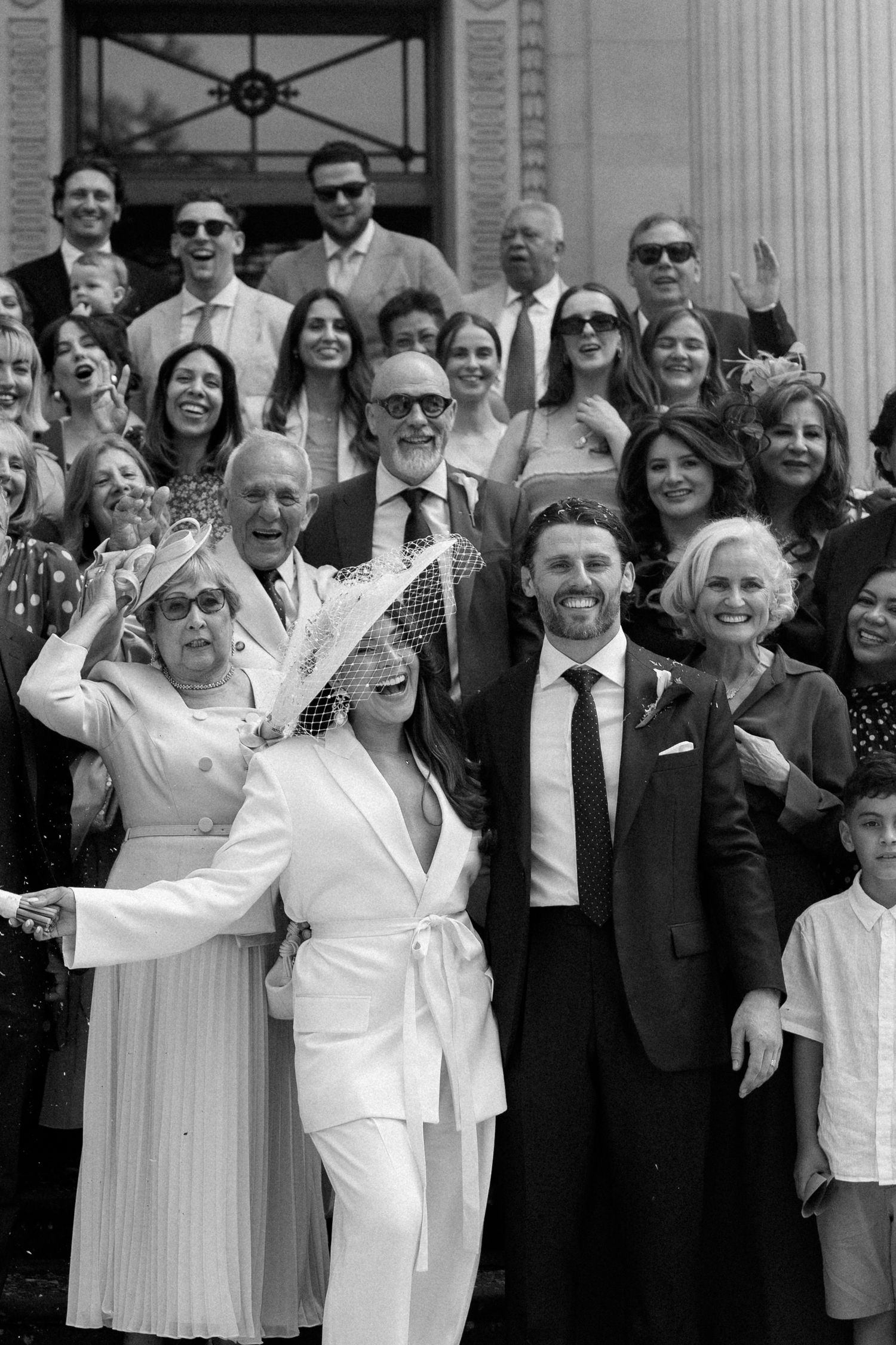 Black and white wedding photo of smiling couple in white suit and dark tuxedo surrounded by joyful guests on steps.