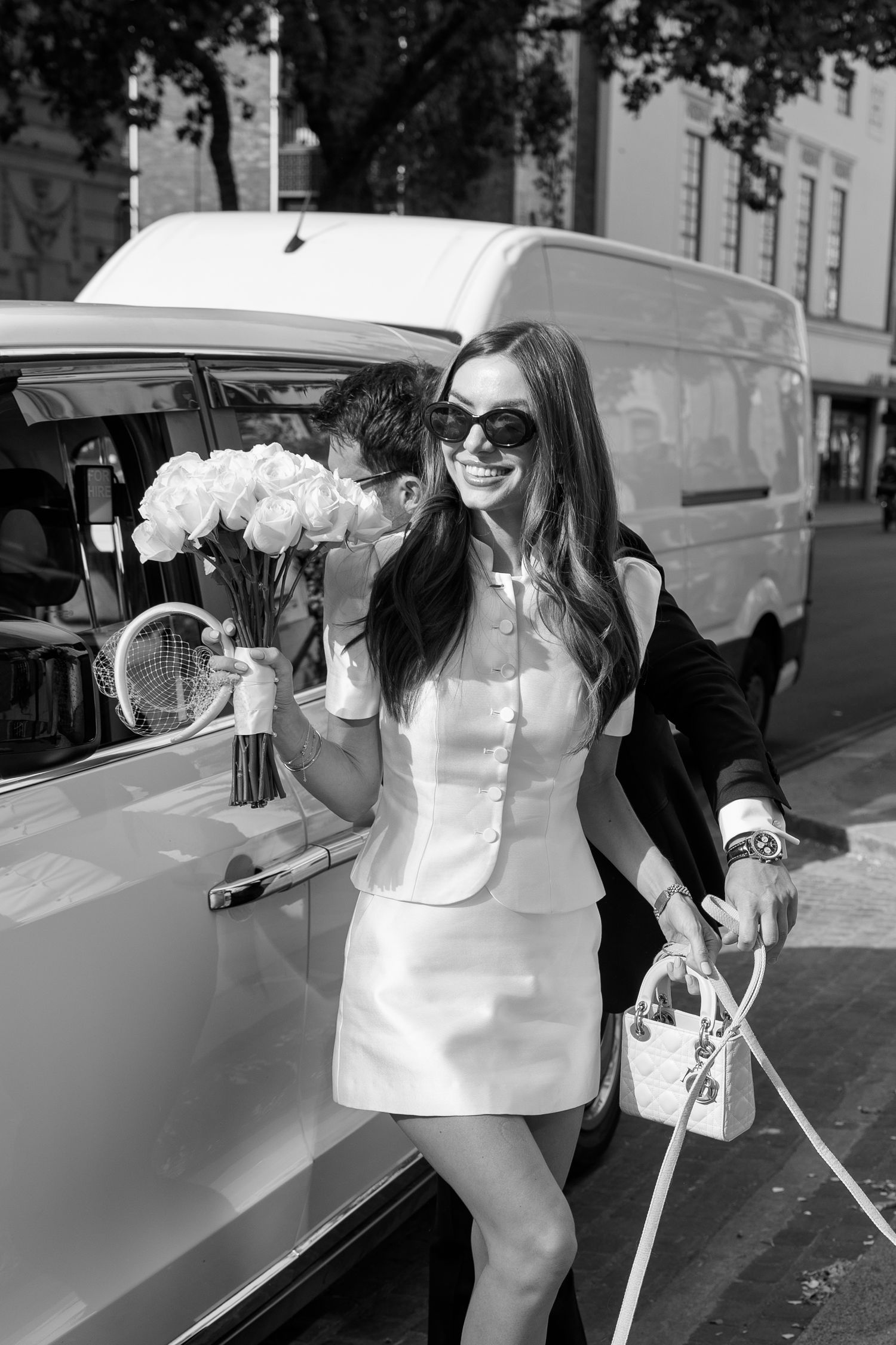 Black and white photo of a smiling woman in white dress holding roses and handbag, exiting a vintage van.