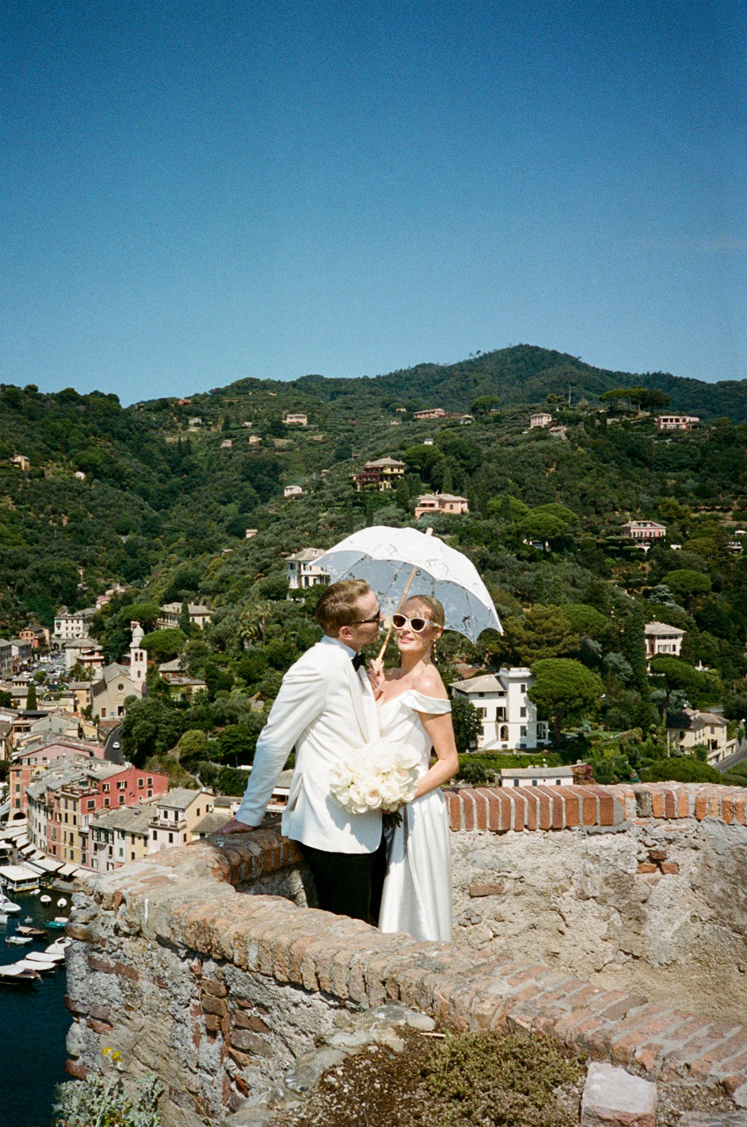 Couple in wedding attire sharing a kiss on a stone wall overlooking Portofino harbor, Italy.