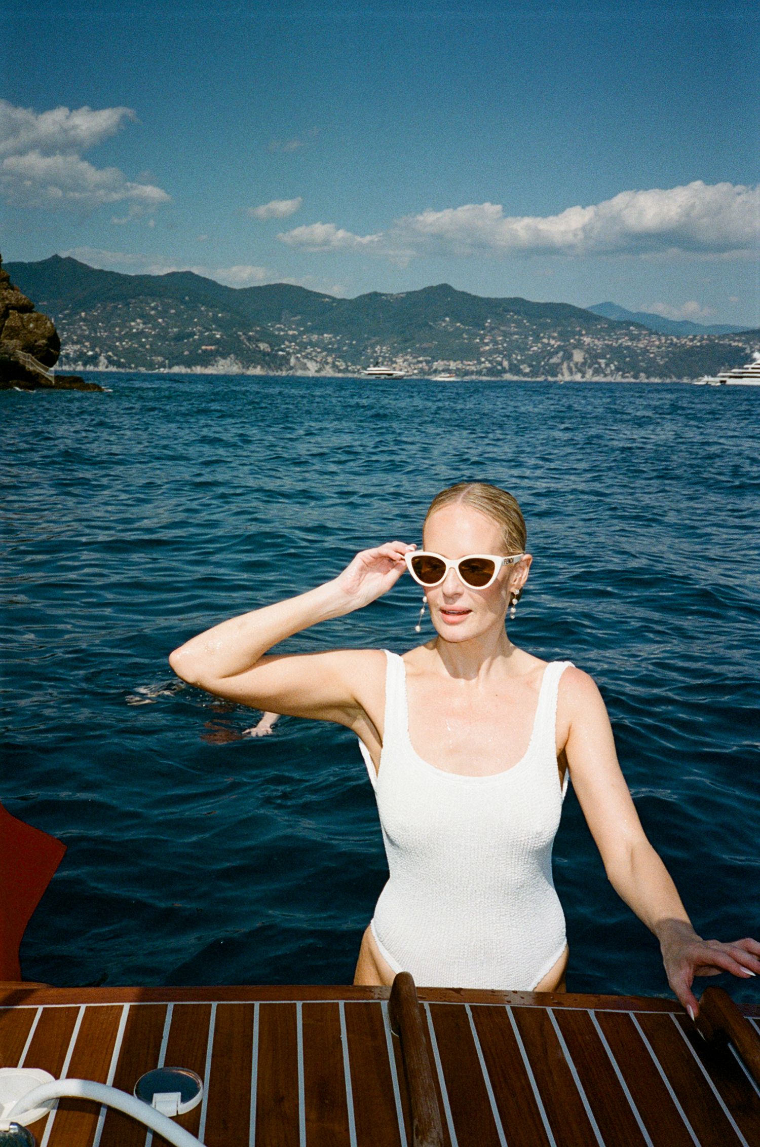 Blonde woman in white swimsuit and cat-eye sunglasses posing on a wooden boat with Italian coastal mountains behind.