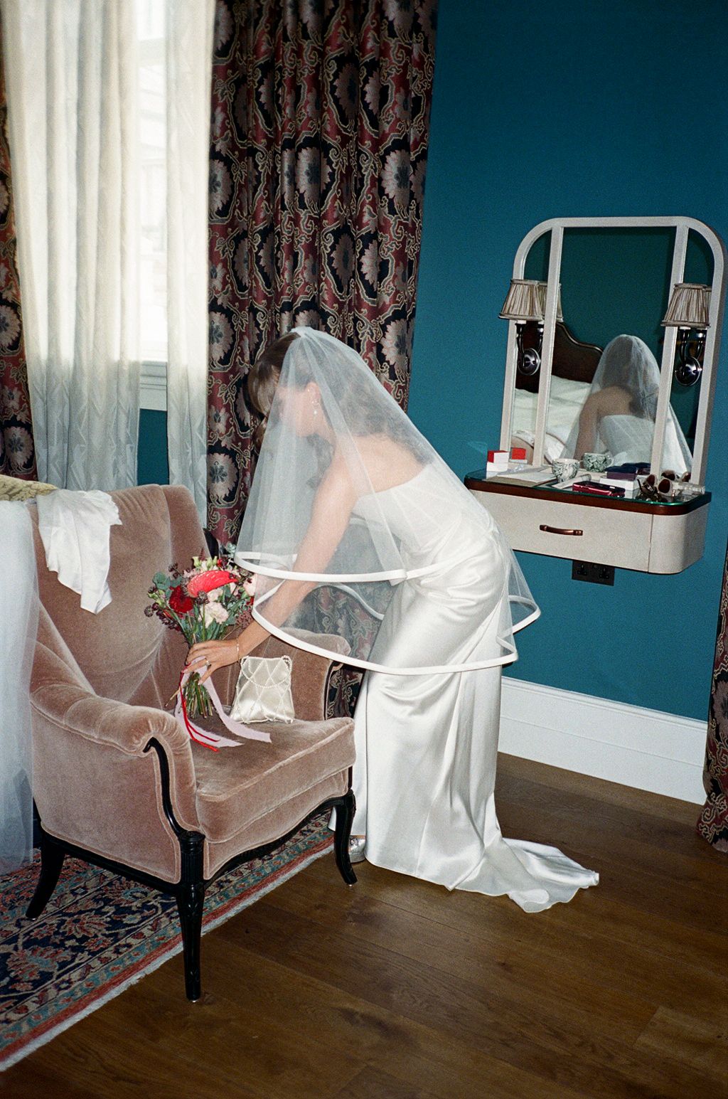 Bride in white satin gown and veil bends over velvet chair holding red and pink bouquet in elegant teal room.